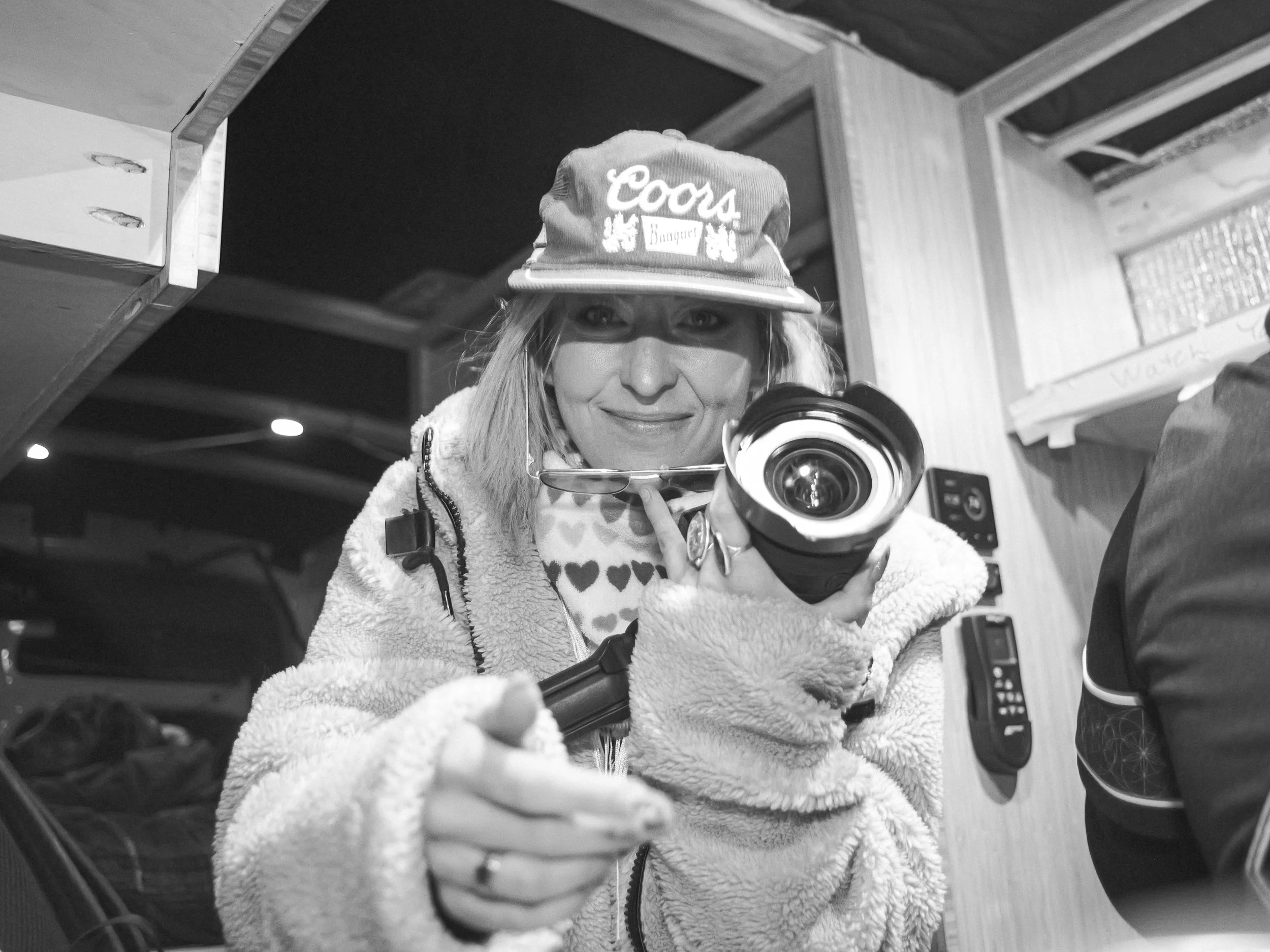 Woman taking a photo with a camera, wearing a cap with 'Coors' and 'Banquet' logos, glasses hanging around her neck, smiling, in a cozy indoor setting.