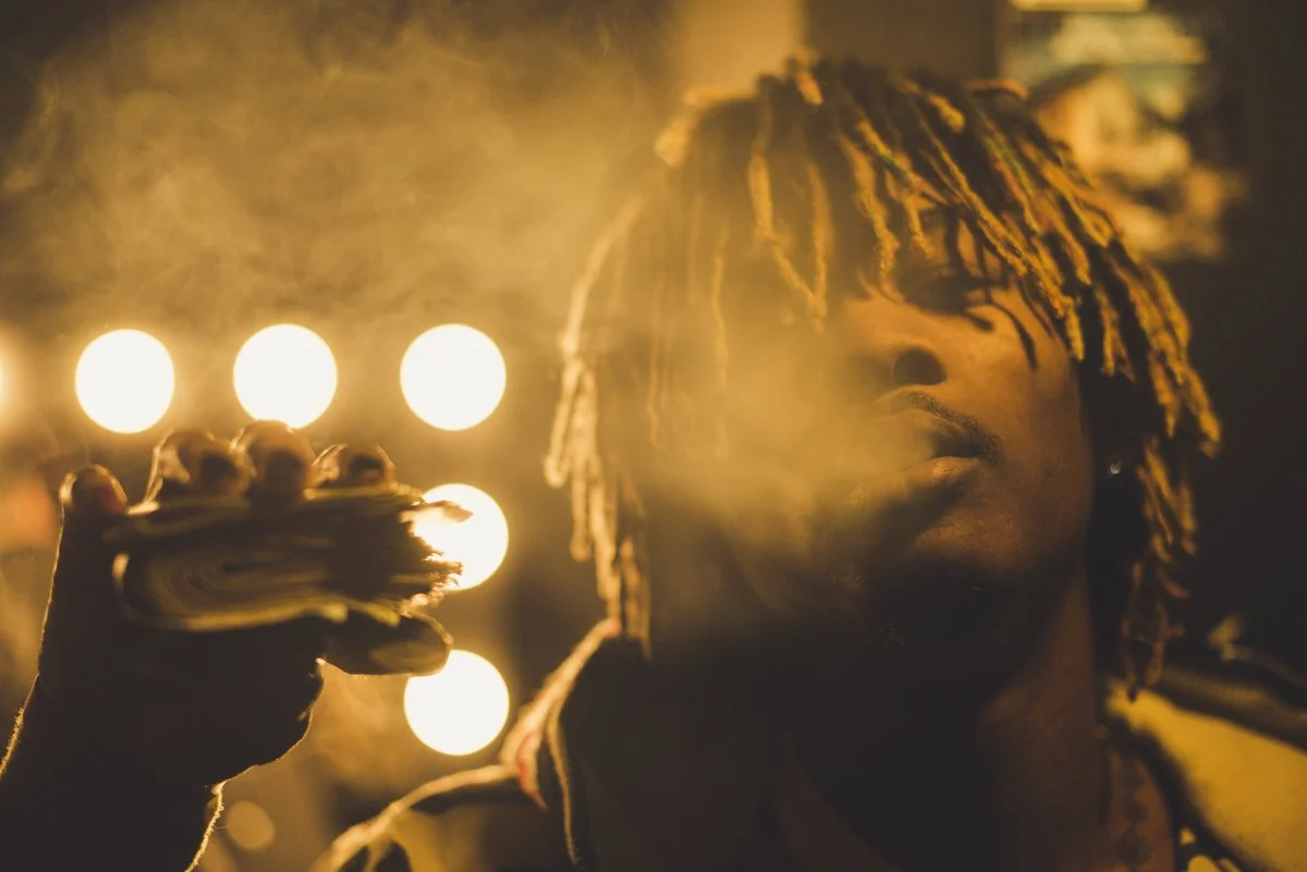 A young man with dreadlocks smoking, with bright warm lights in the background creating a hazy atmosphere.
