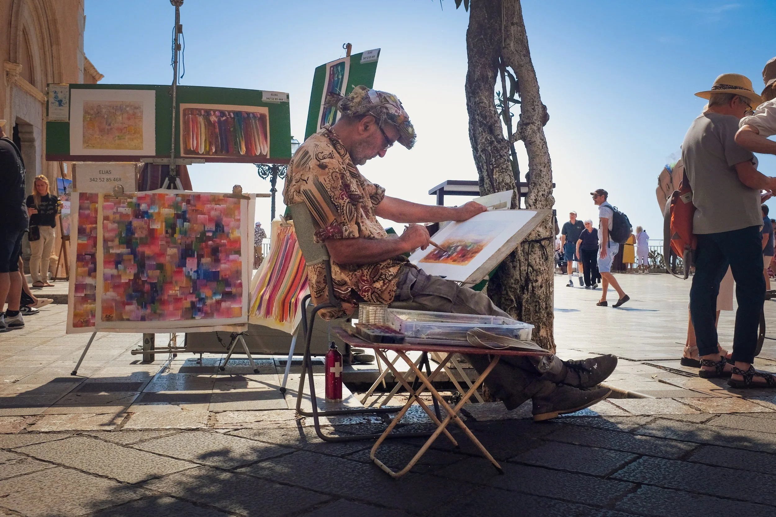 Street artist sitting on a folding chair, painting on a canvas with colorful abstract art, surrounded by paintings and art supplies, with pedestrians walking by on a sunny day.