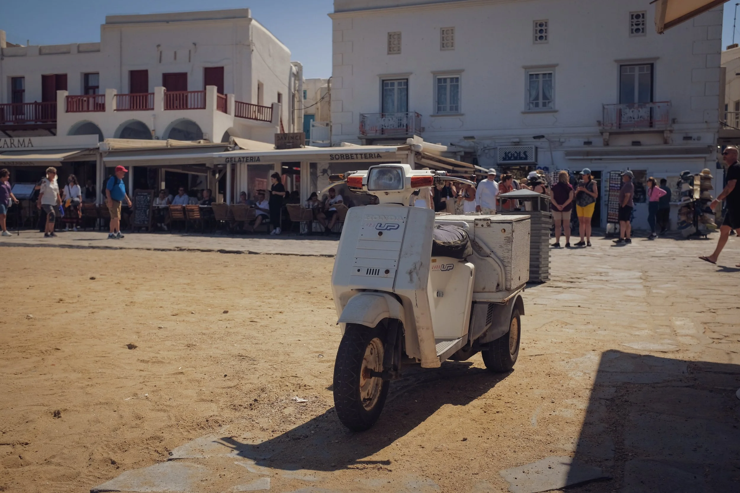 An old white scooter parked on sandy ground in front of a busy outdoor cafe with people walking and dining in a Mediterranean town.