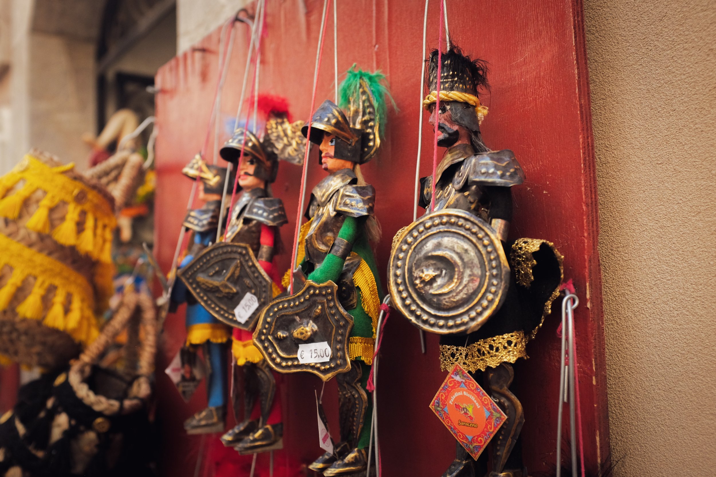 Colorful handcrafted wooden puppets dressed as medieval soldiers with shields and helmets, hanging on a red display wall in a market.