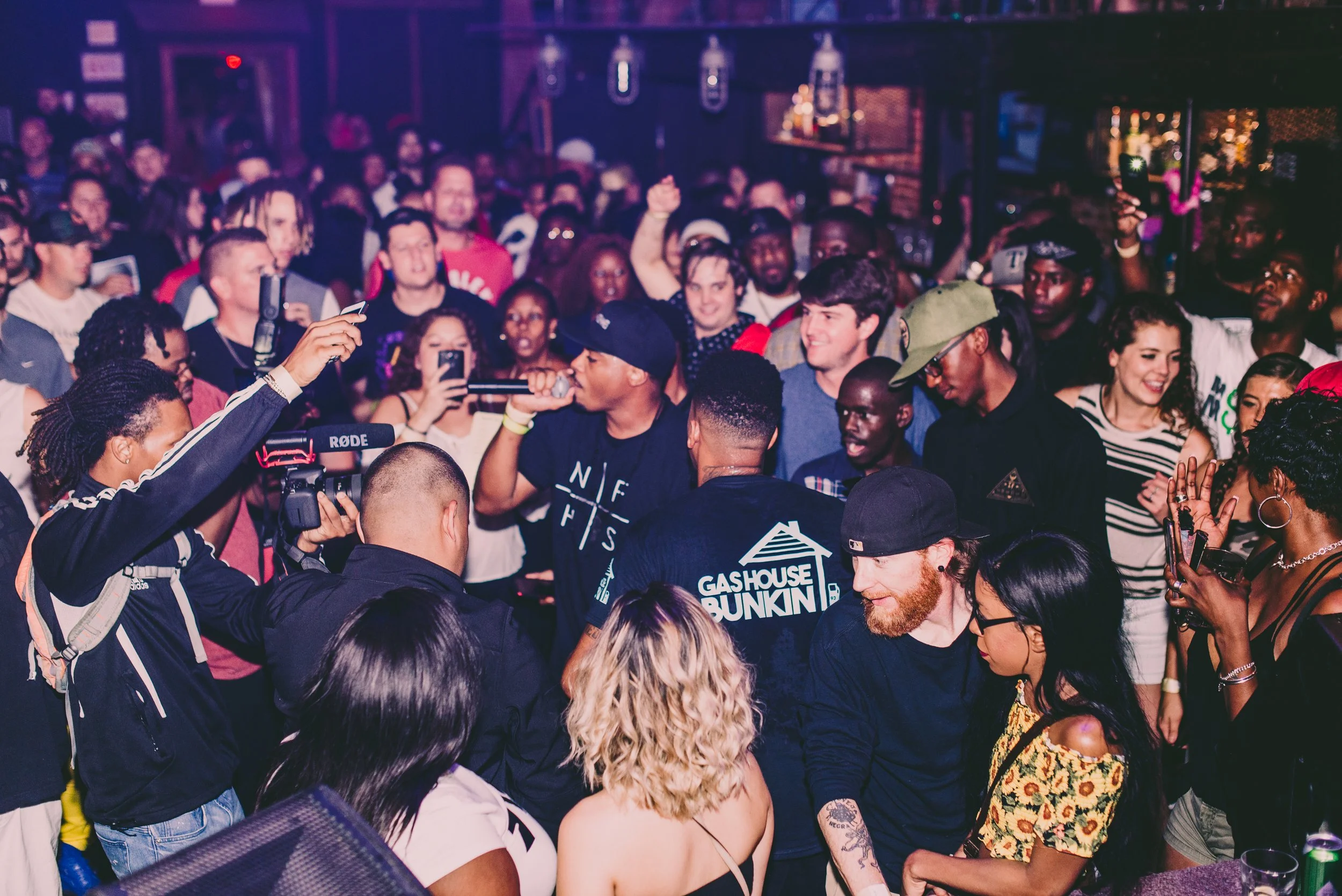 A crowd of people at a lively indoor concert or event, with a man in a black t-shirt singing into a microphone, surrounded by friends and fans taking photos and watching.