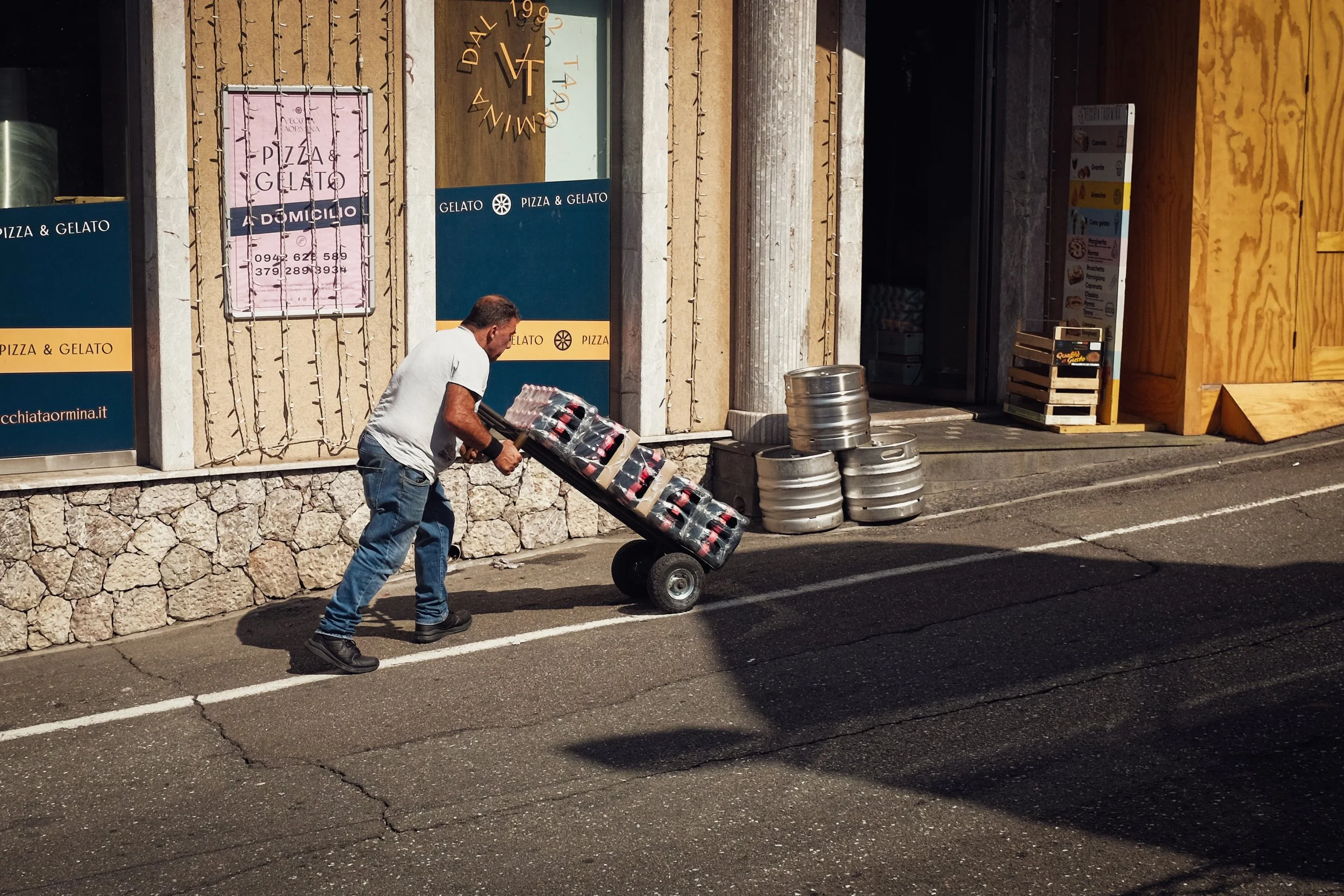 A man pushing a hand truck loaded with bottles outside a building with signs advertising pizza and gelato, and stacks of aluminum beer kegs on the sidewalk.
