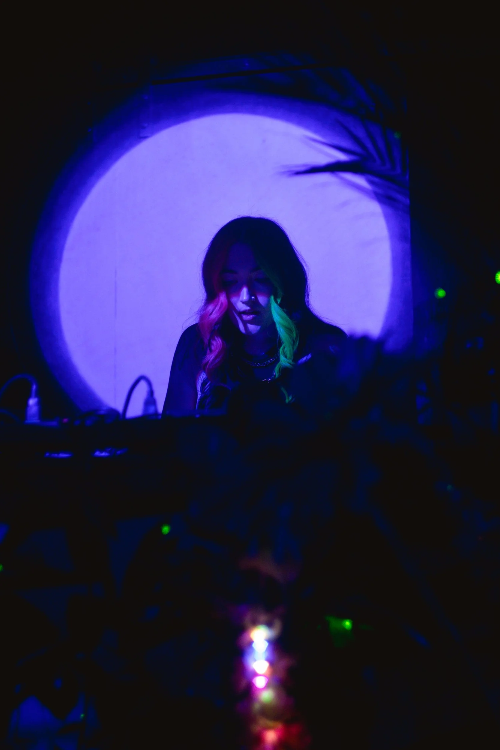 A woman with long hair, partly neon pink and partly neon green, is DJing in a dark room illuminated by blue and purple lights. A large full moon projection is in the background.