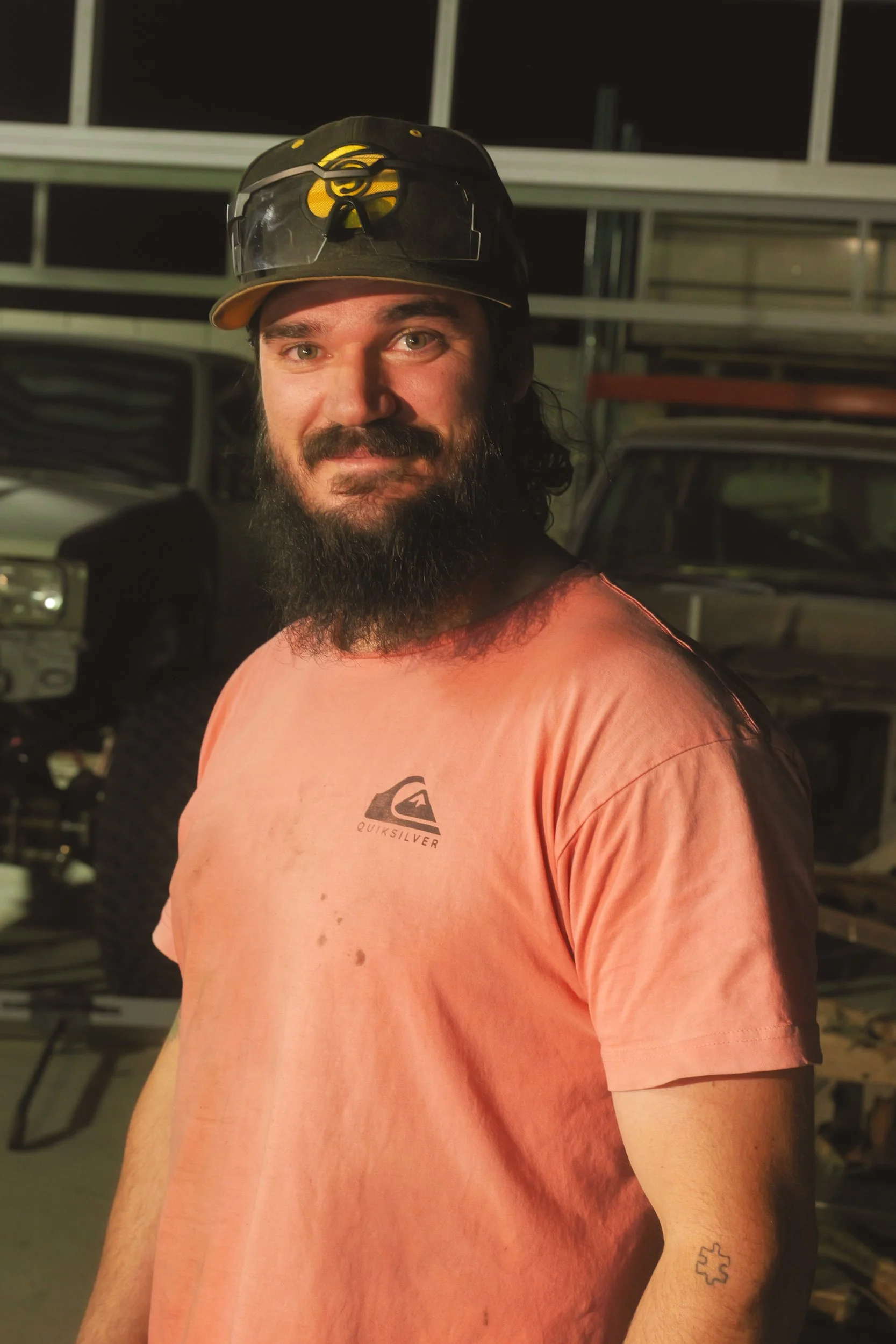 Man with a beard wearing a black cap with sunglasses on top, an orange Quiksilver t-shirt, and a tattoo on his left arm, standing in a garage with vehicles in the background.