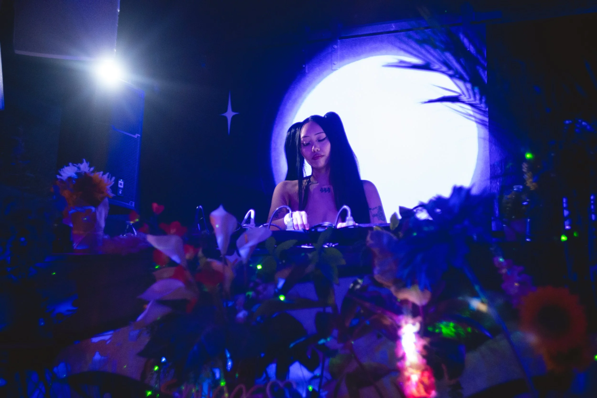 A female DJ with long black hair styled in pigtails performs at a nightclub with a bright full moon backdrop and colorful flowers around the DJ booth.