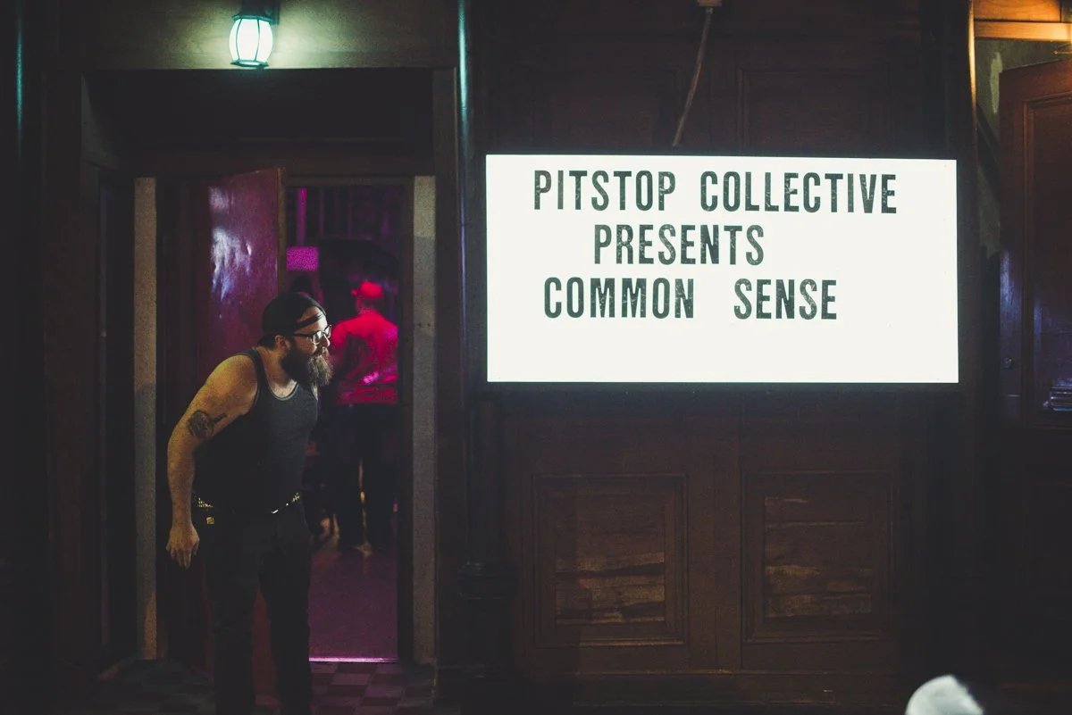 A man with a beard and glasses, wearing a black tank top and beanie, looks at a lit sign that reads 'Pitstop Collective Presents Common Sense' in a dimly lit room with dark wood paneling, as other people are visible in the background.