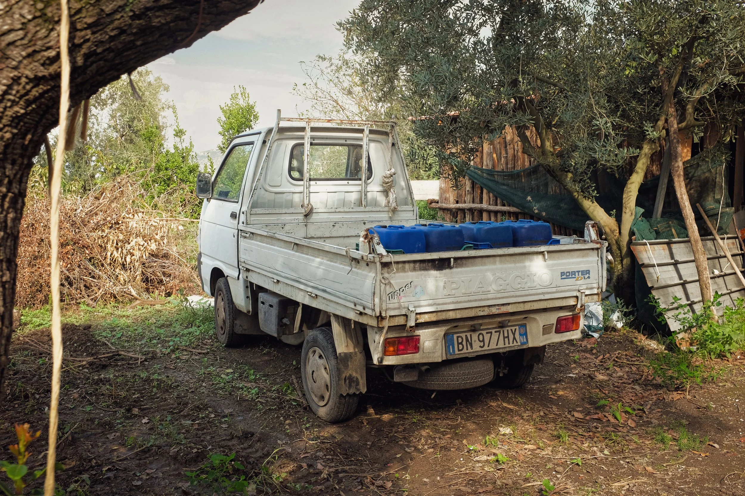 A small white pickup truck with blue containers in the bed, parked on dirt ground near trees and a wooden shed.