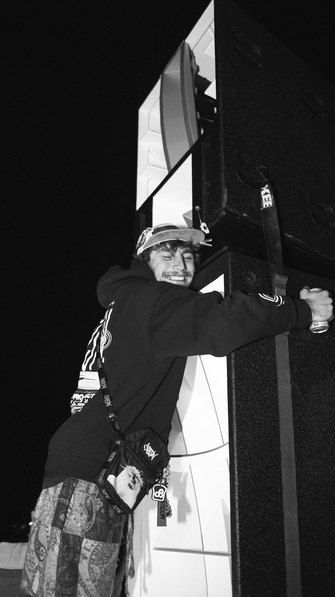A young man with a big smile hugging a large speaker or audio equipment at night, wearing a hoodie, patterned pants, a cap, and a crossbody bag with various patches and accessories.