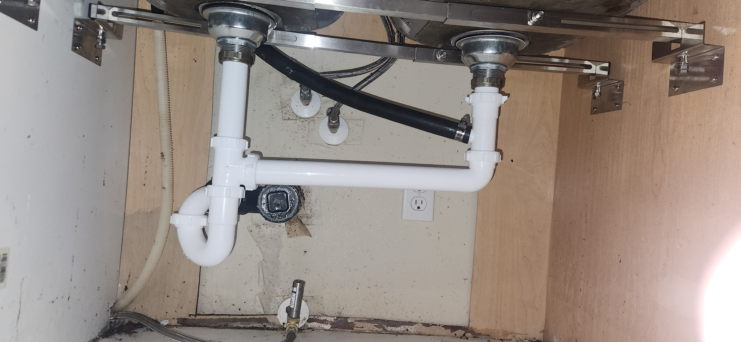 Under-sink plumbing with white PVC pipes, black flexible hose, and metal fittings, mounted on a wall with a wooden countertop and an electrical outlet nearby.