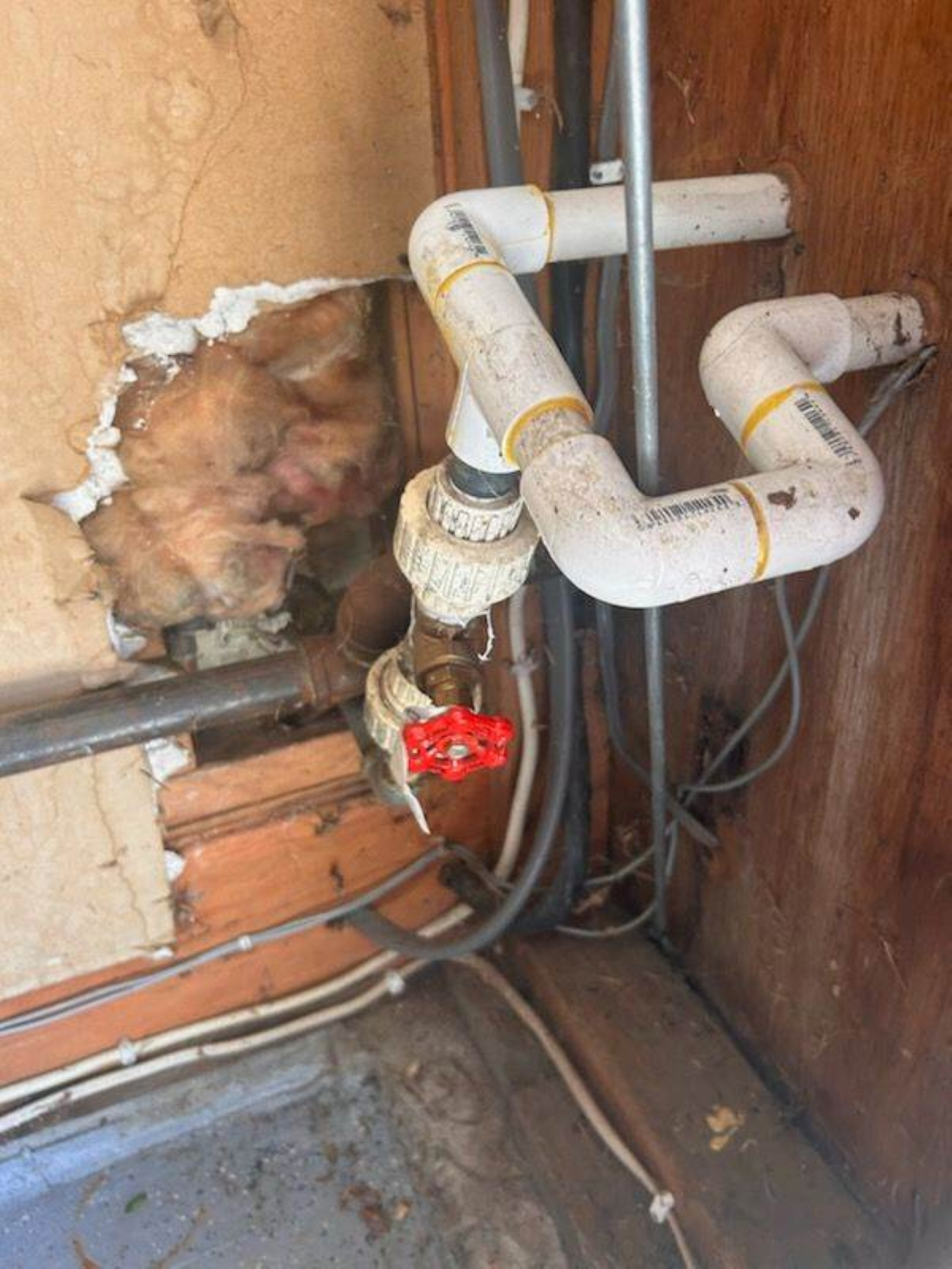 A person looking through a hole in a wall at plumbing pipes and a red valve handle.