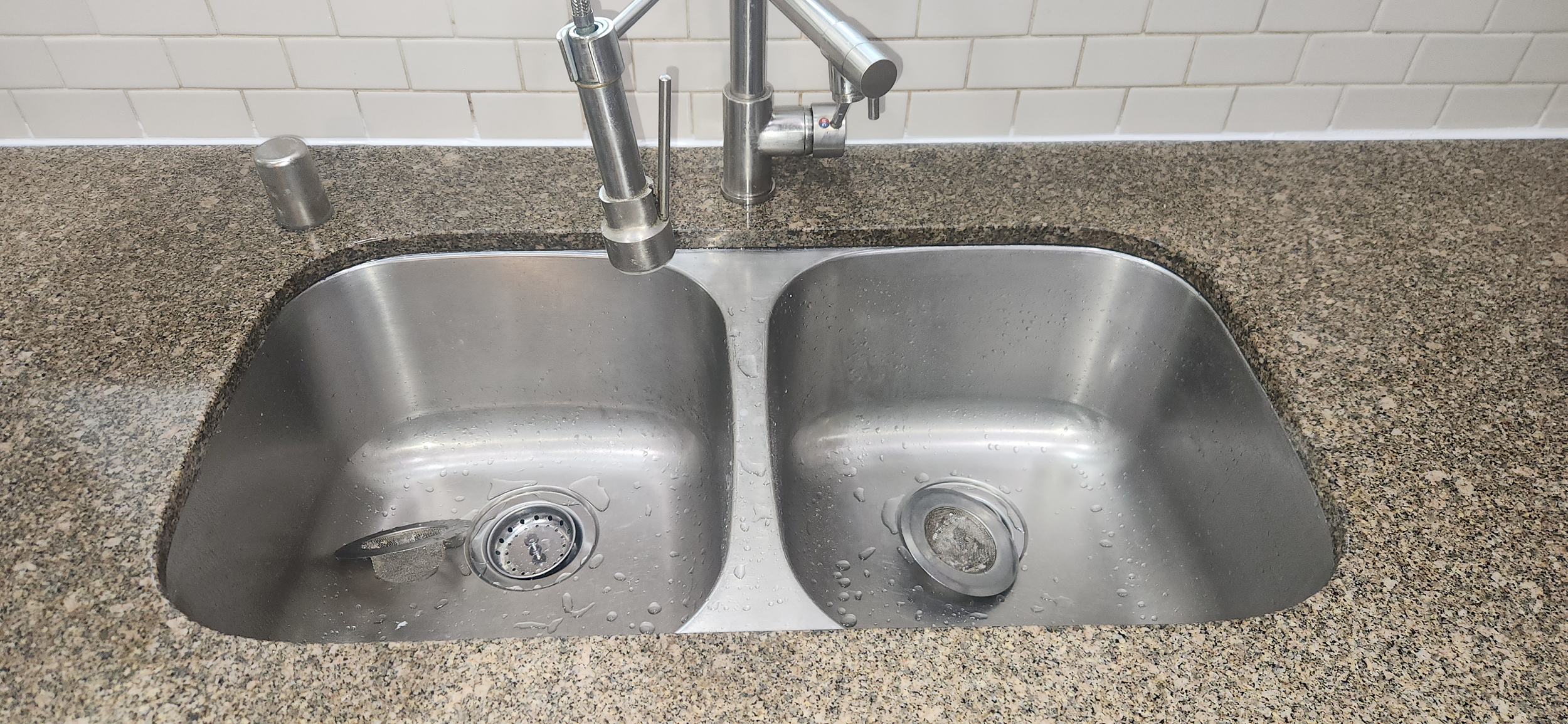Stainless steel kitchen sink with two basins, water droplets inside, and a modern dual-handle faucet, set in a speckled granite countertop with a white tile backsplash.