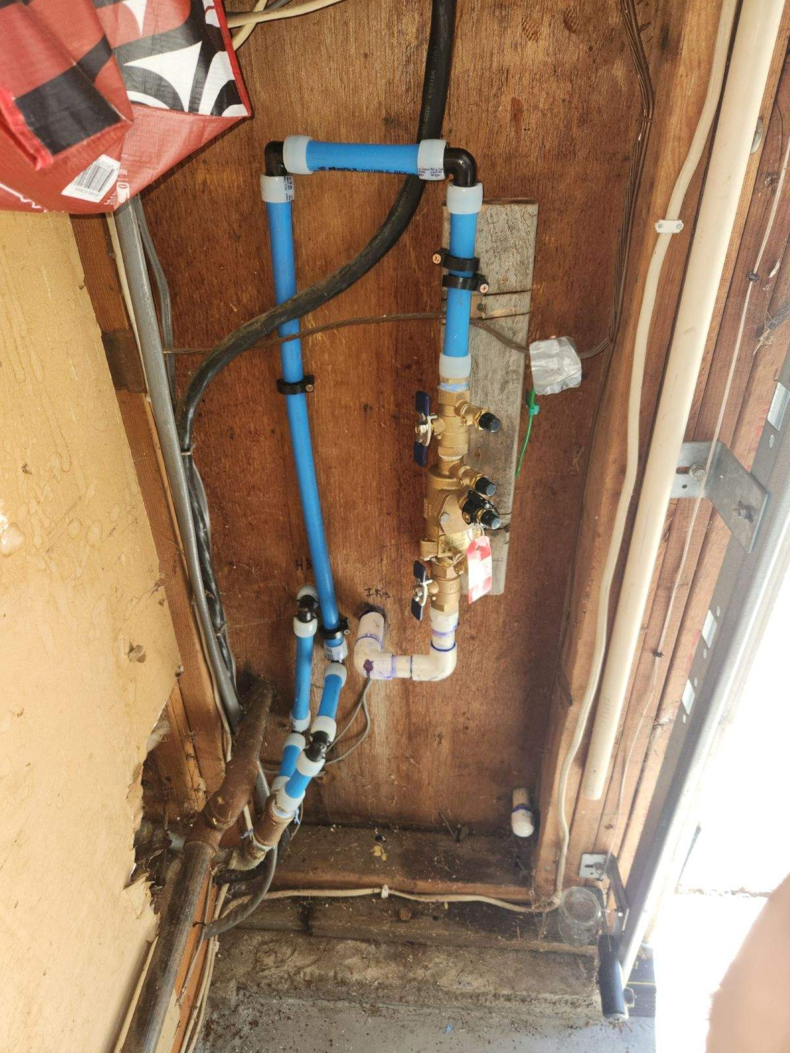 Plumbing pipes and a brass valve assembly mounted on wooden wall in a basement or utility room.
