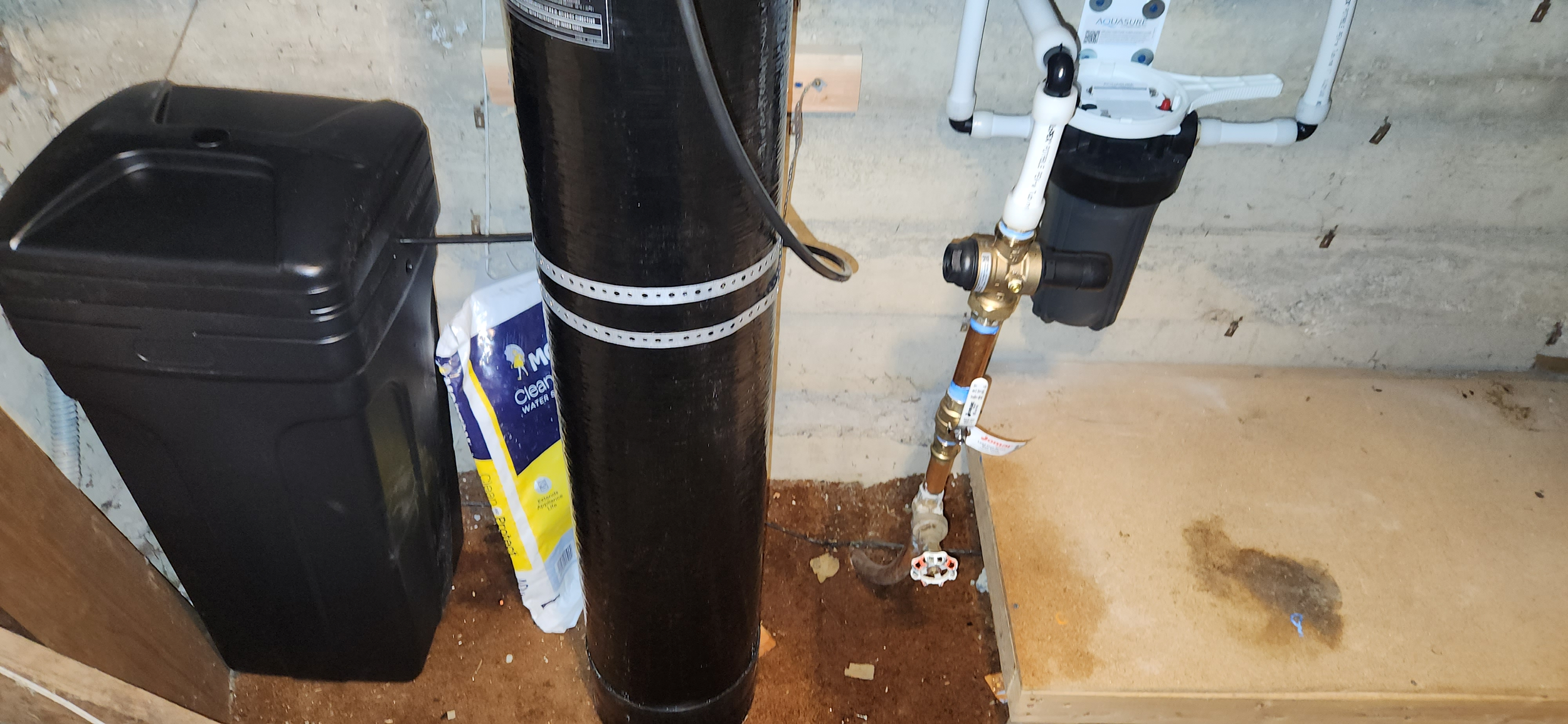 A black water softener or filtration unit with a large black pipe next to it, and a plumbing system with PVC pipes and a brass valve mounted on a concrete wall inside a basement or utility room.