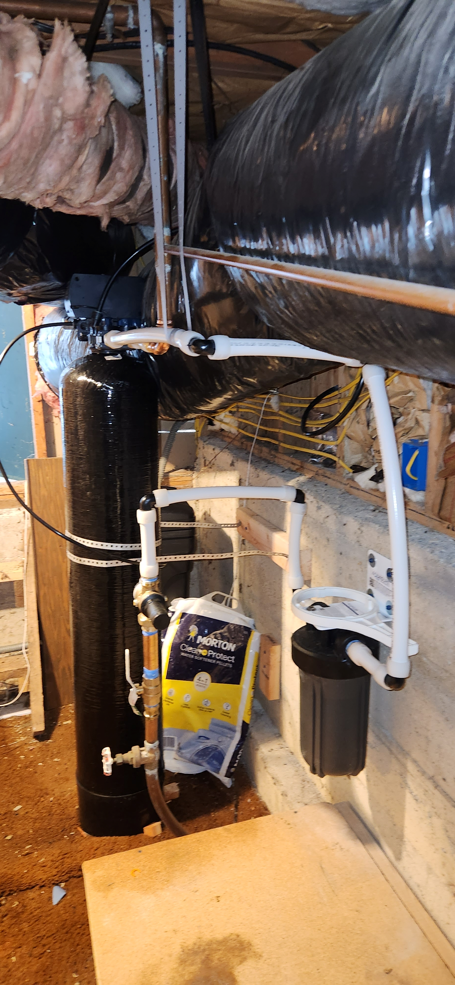 A basement water filtration system installed against a cinderblock wall, with pipes, filters, and a black tank, in a room with unfinished walls and exposed wood beams.