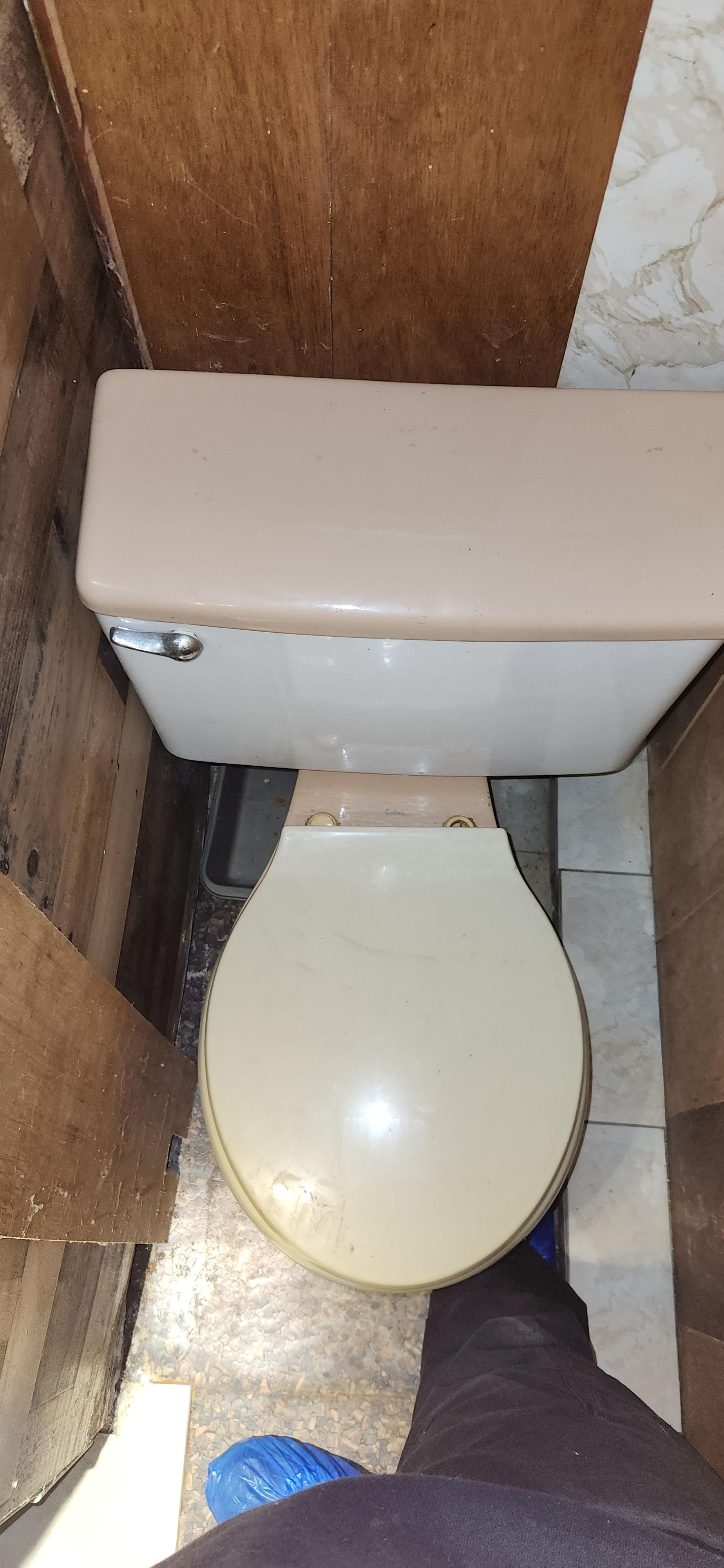 Overhead view of an old toilet with a beige seat and tank, positioned between wooden and tiled walls, with a person's foot visible in the bottom right corner.