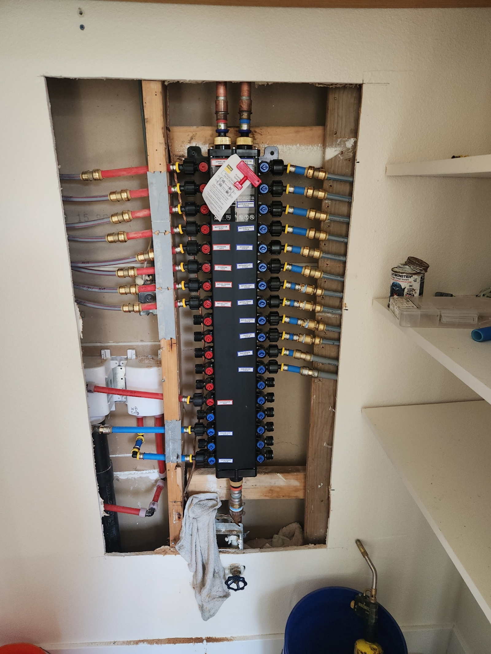 A plumbing manifold with multiple hot and cold water supply lines connected, set in a wall with shelves on the right side.