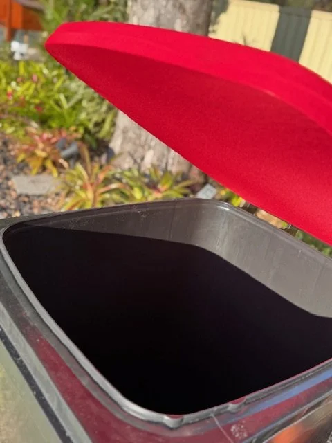 Empty trash bin with red lid open, outdoors with plants and a wooden fence in the background.