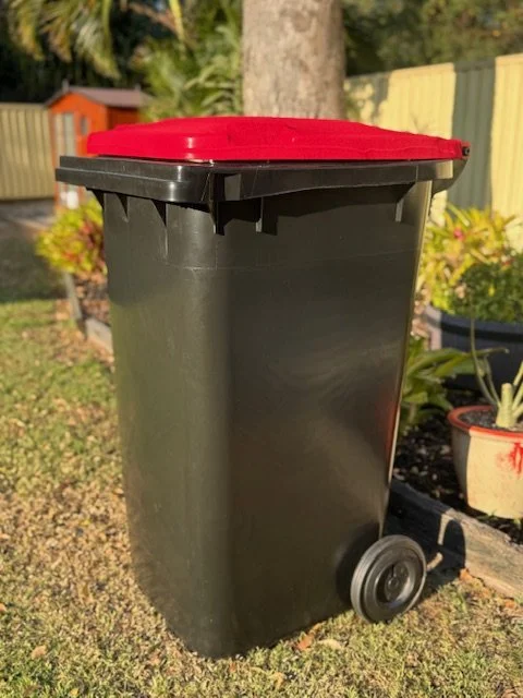 A black outdoor trash can with a red lid on a grassy area.