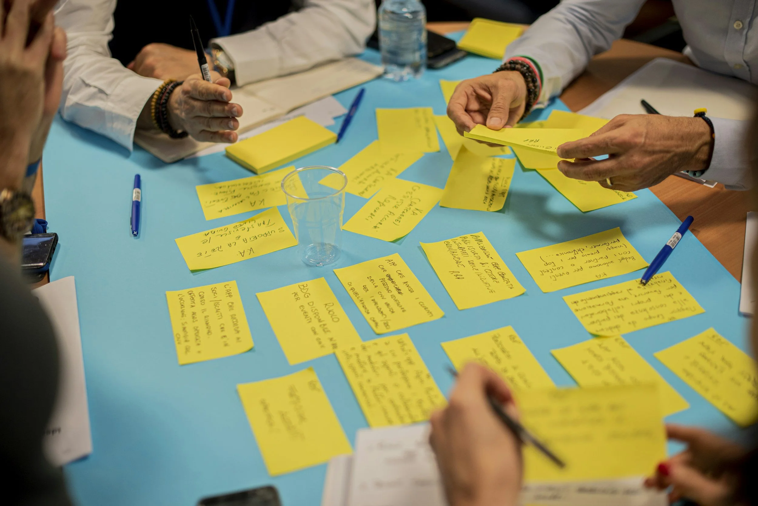 People participating in a brainstorming session, writing on and organizing yellow sticky notes on a blue table, with pens, notebooks, and water bottles visible.