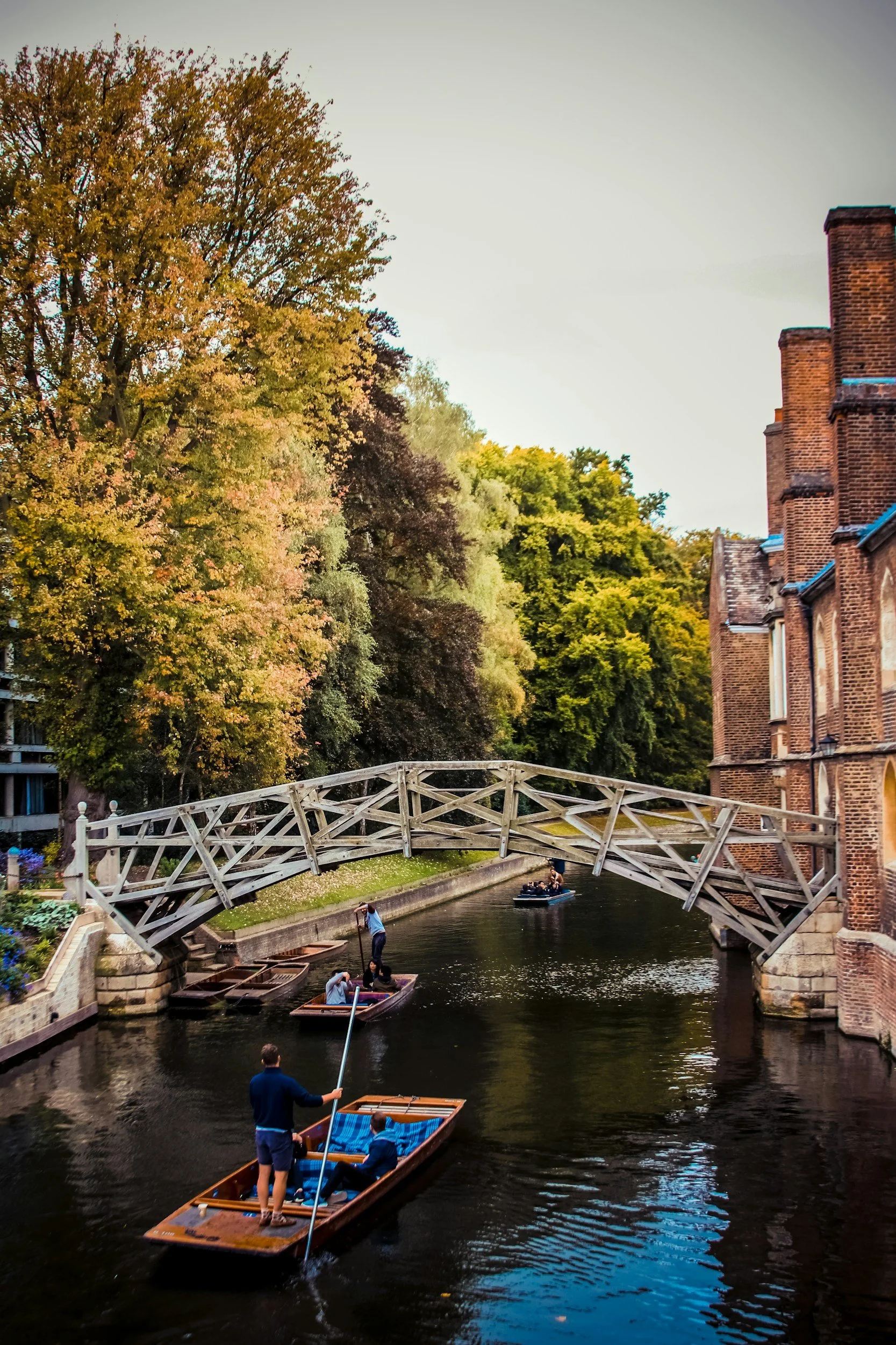 People enjoying boat rides under a small wooden bridge on a narrow canal, with tall trees and brick buildings on the sides.
