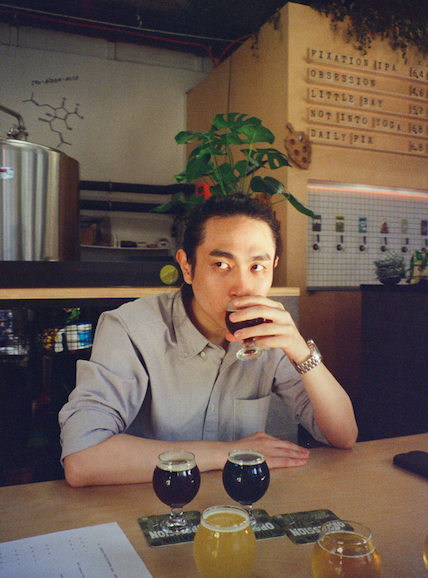 A person sitting at a bar or restaurant table, drinking a dark beverage from a glass, with three other similar drinks and a menu in front. The background features a plant, a chalkboard, and a sign with menu and pricing info.
