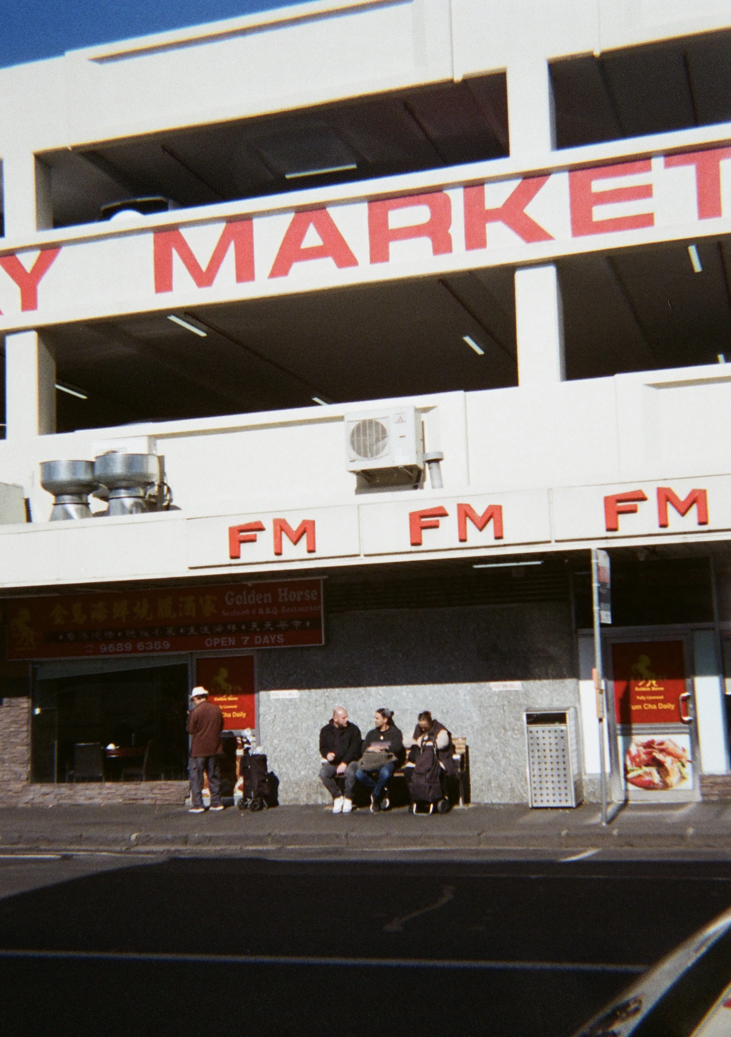 People sitting and standing outside a restaurant under a building with large red signs that say 'FM' and a partial sign with 'MARKET' in red, and an air conditioning unit on the second floor.