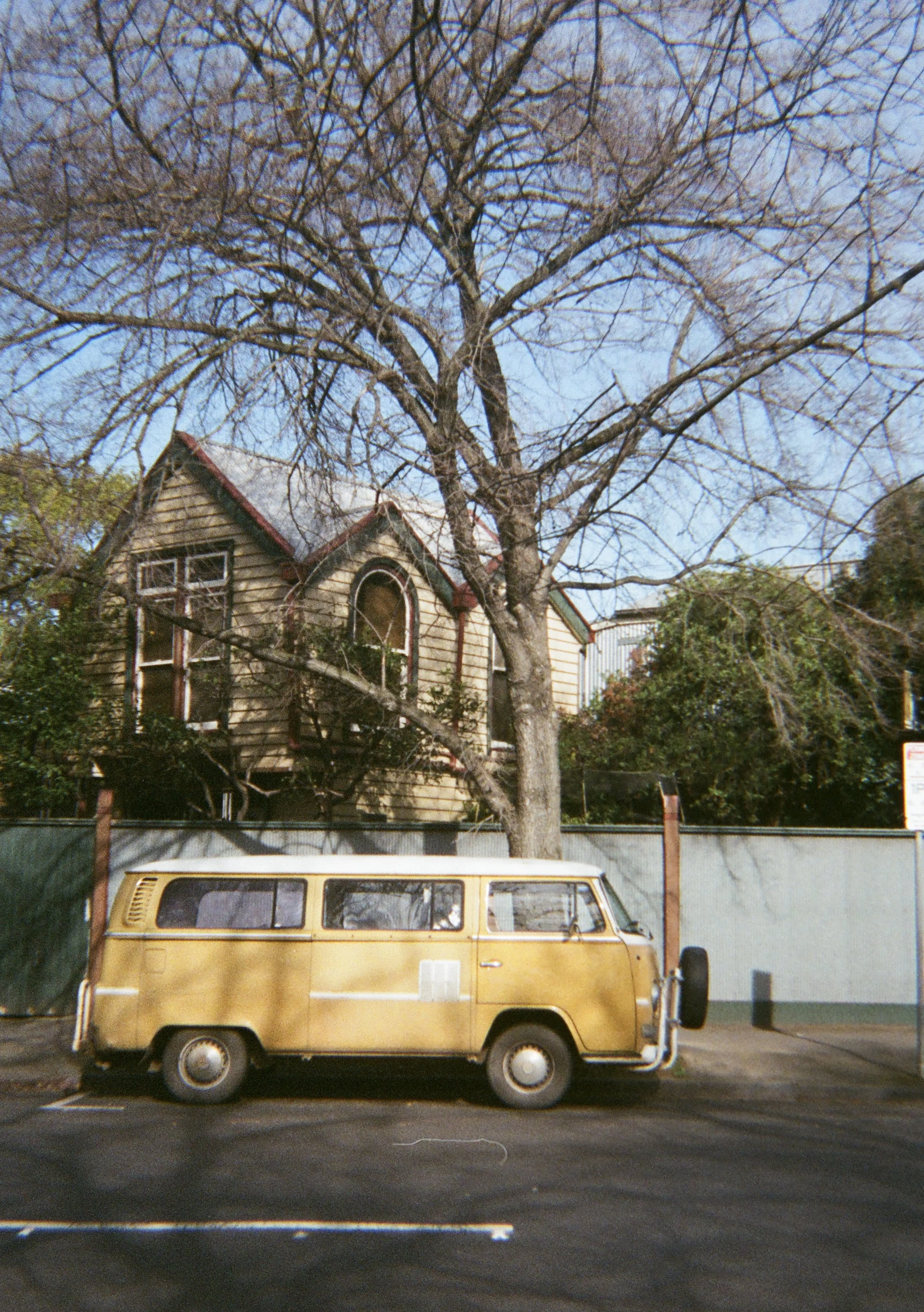 A yellow vintage minivan parked in front of a house with a large leafless tree in the yard during daytime.
