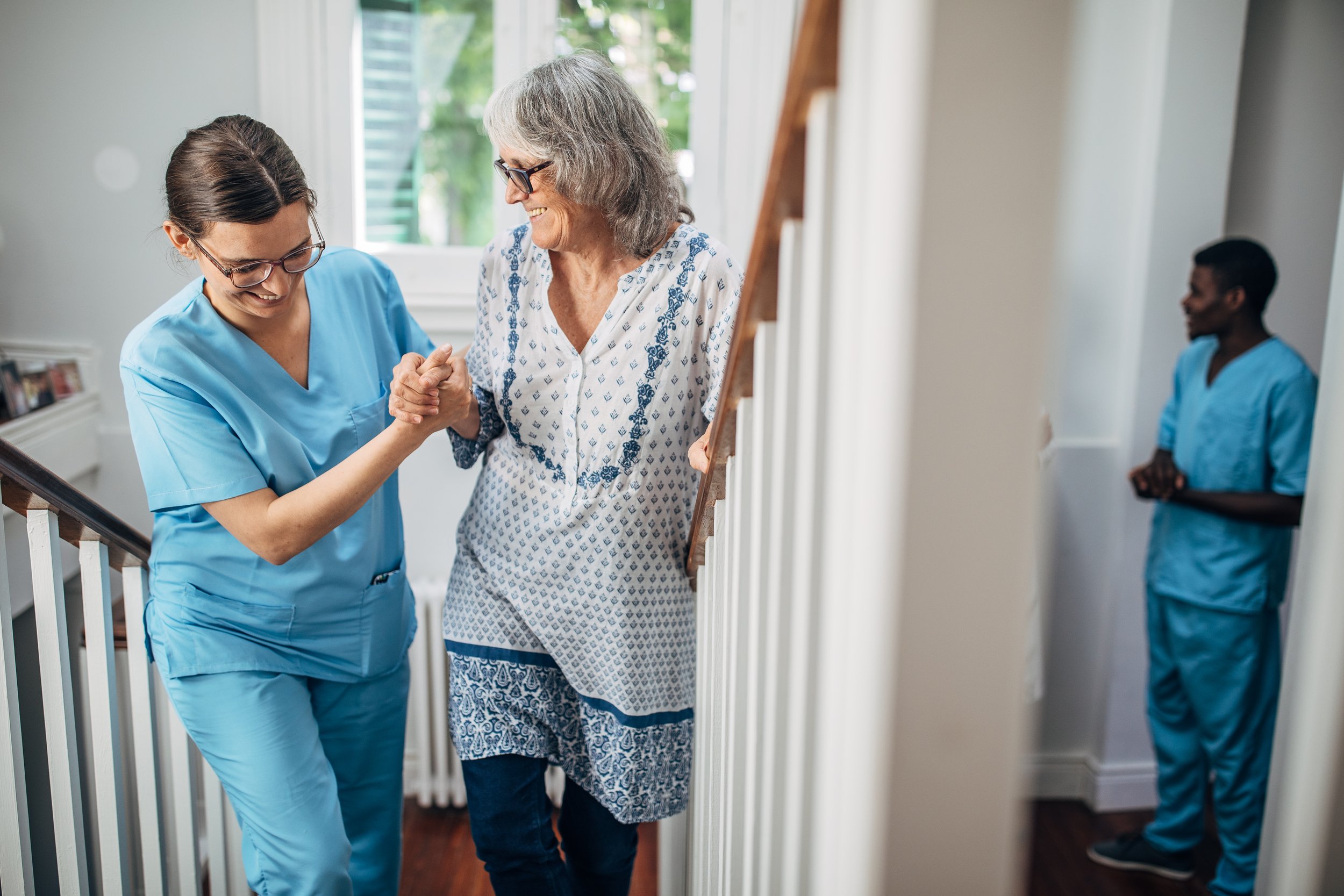 A nurse helping an elderly woman walk up the stairs inside a home, with a second nurse standing in the background.