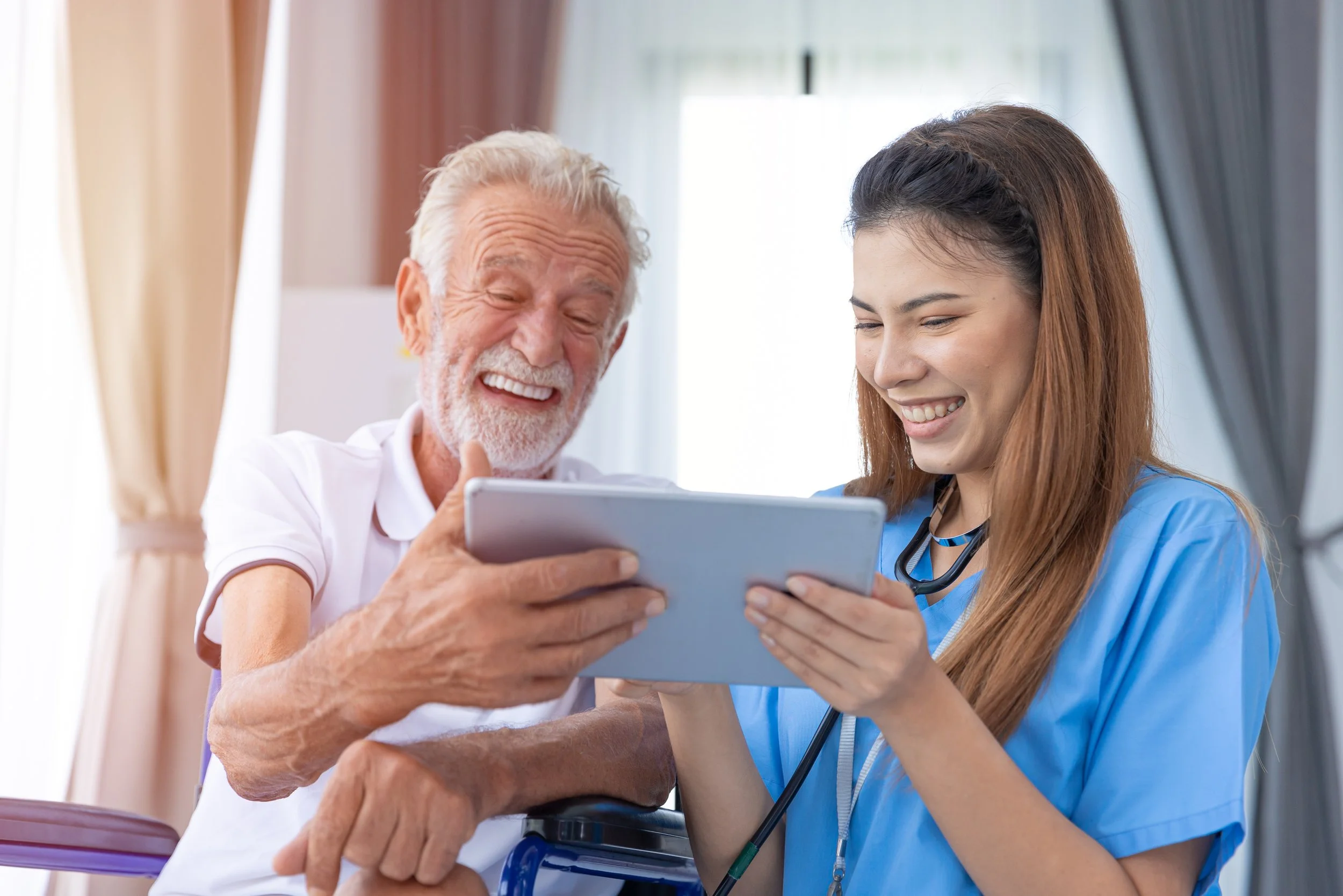 An elderly man and a young female nurse are smiling and looking at a tablet together in a bright room.