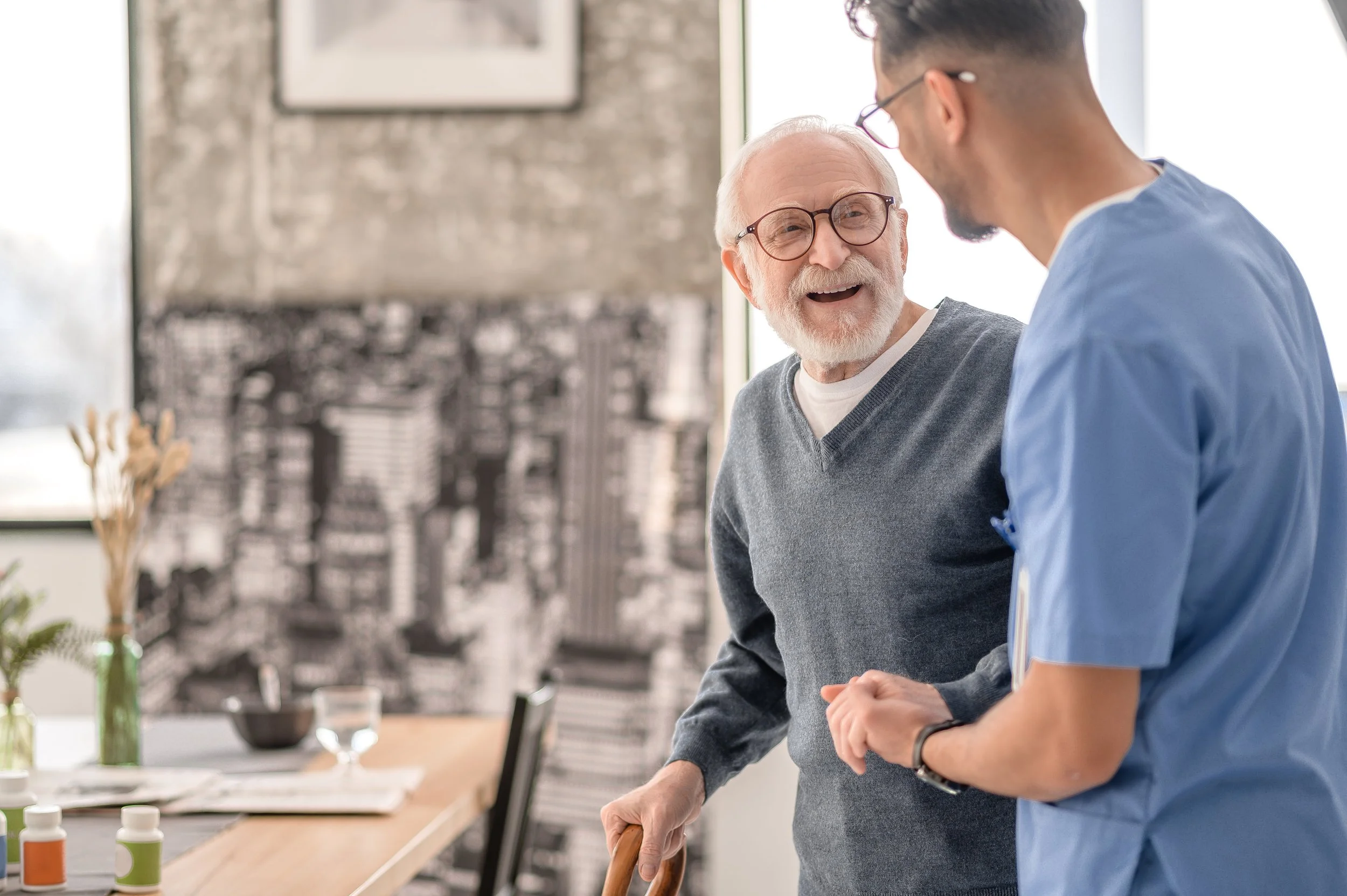 An elderly man with glasses and a gray sweater smiling and talking with a young male healthcare professional in a blue uniform in a bright room.