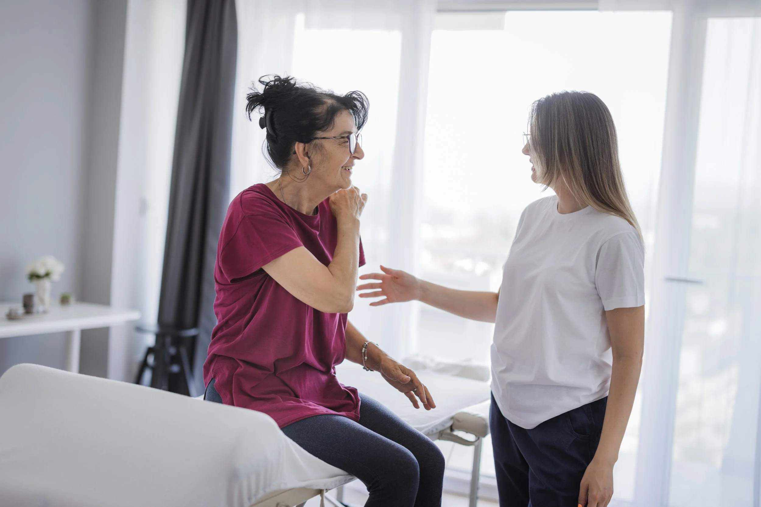 A young woman and an older woman in a bright room, smiling at each other. The older woman is sitting on a medical examination table, wearing a maroon shirt and glasses. The younger woman, standing nearby, is wearing a white t-shirt and dark pants, reaching out toward the older woman as they converse.