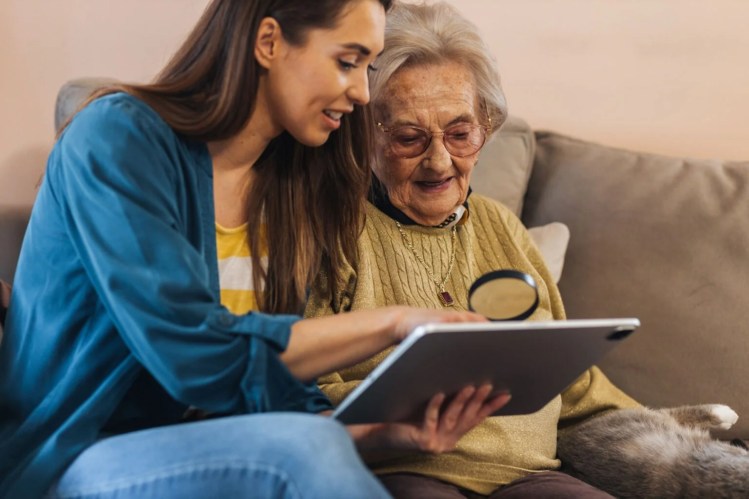 A young woman and an elderly woman sitting on a couch looking at a tablet together, with the young woman showing something to the elderly woman.