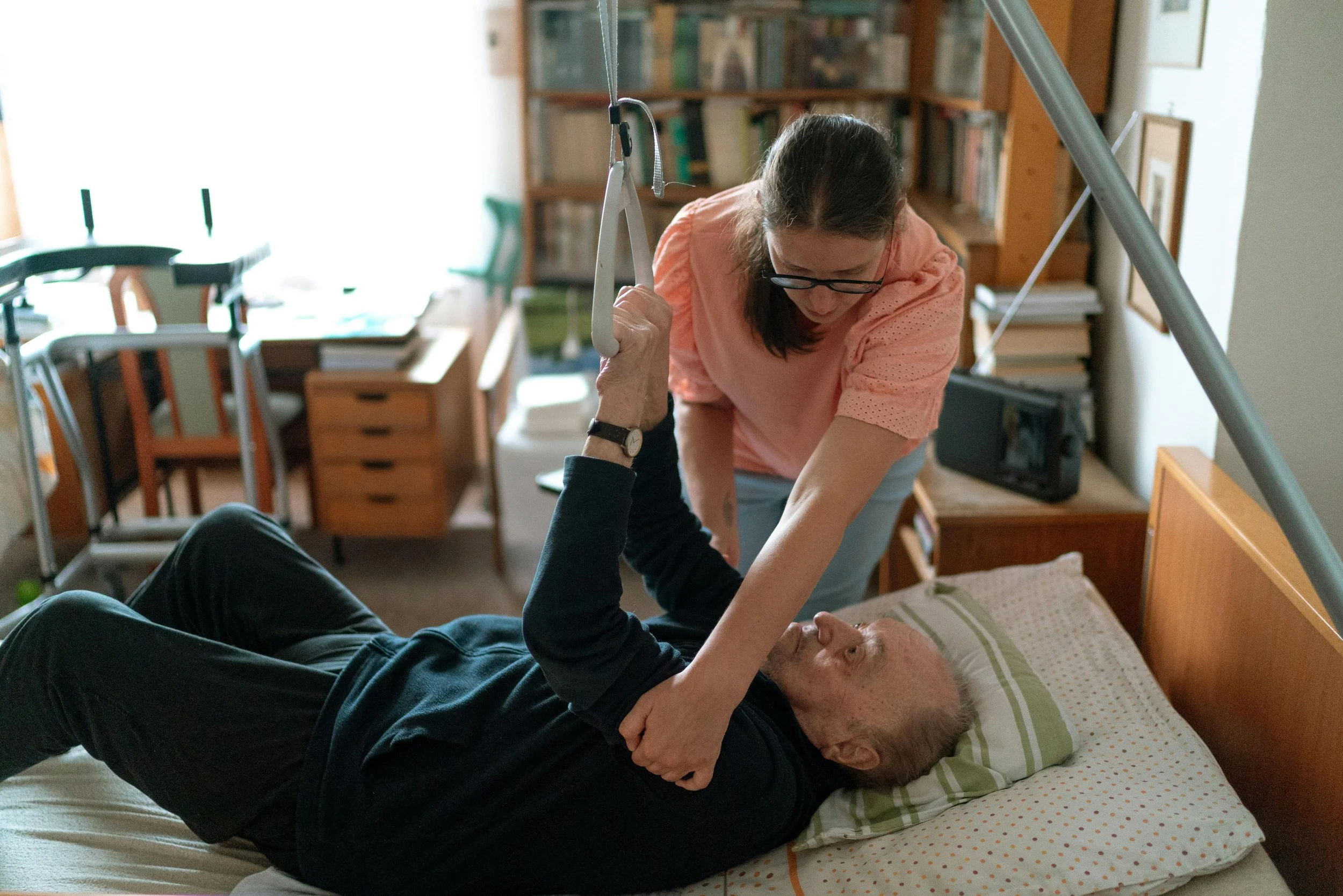 A woman helps an elderly man to transfer from his bed using a transfer sling. The man is lying on his back on the bed, and the woman is standing next to him, holding the sling handles. The scene is set in a room with bookshelves, a window, and various furniture.