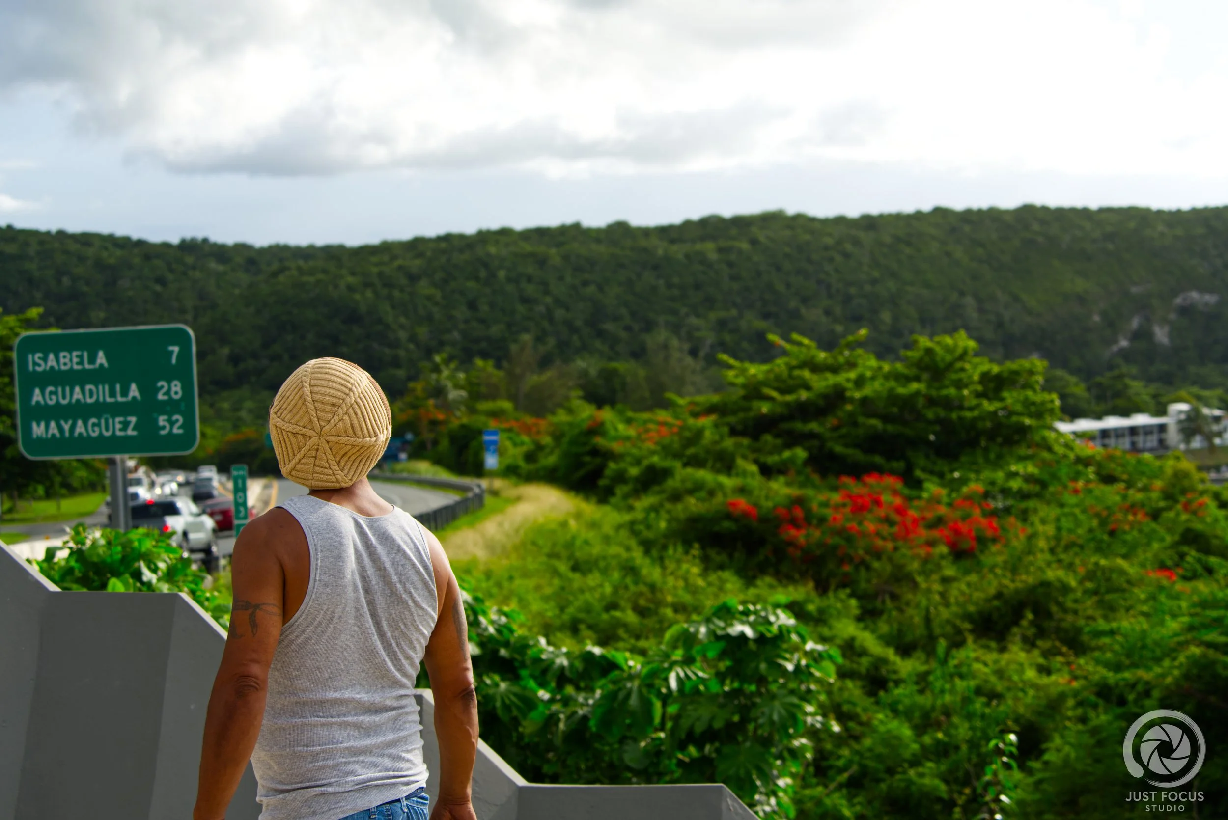 A person with a tan hat and gray shirt standing outdoors near a highway, with green trees, hills, and a road sign indicating directions to Isabel, Aguadilla, and Mayaguez.