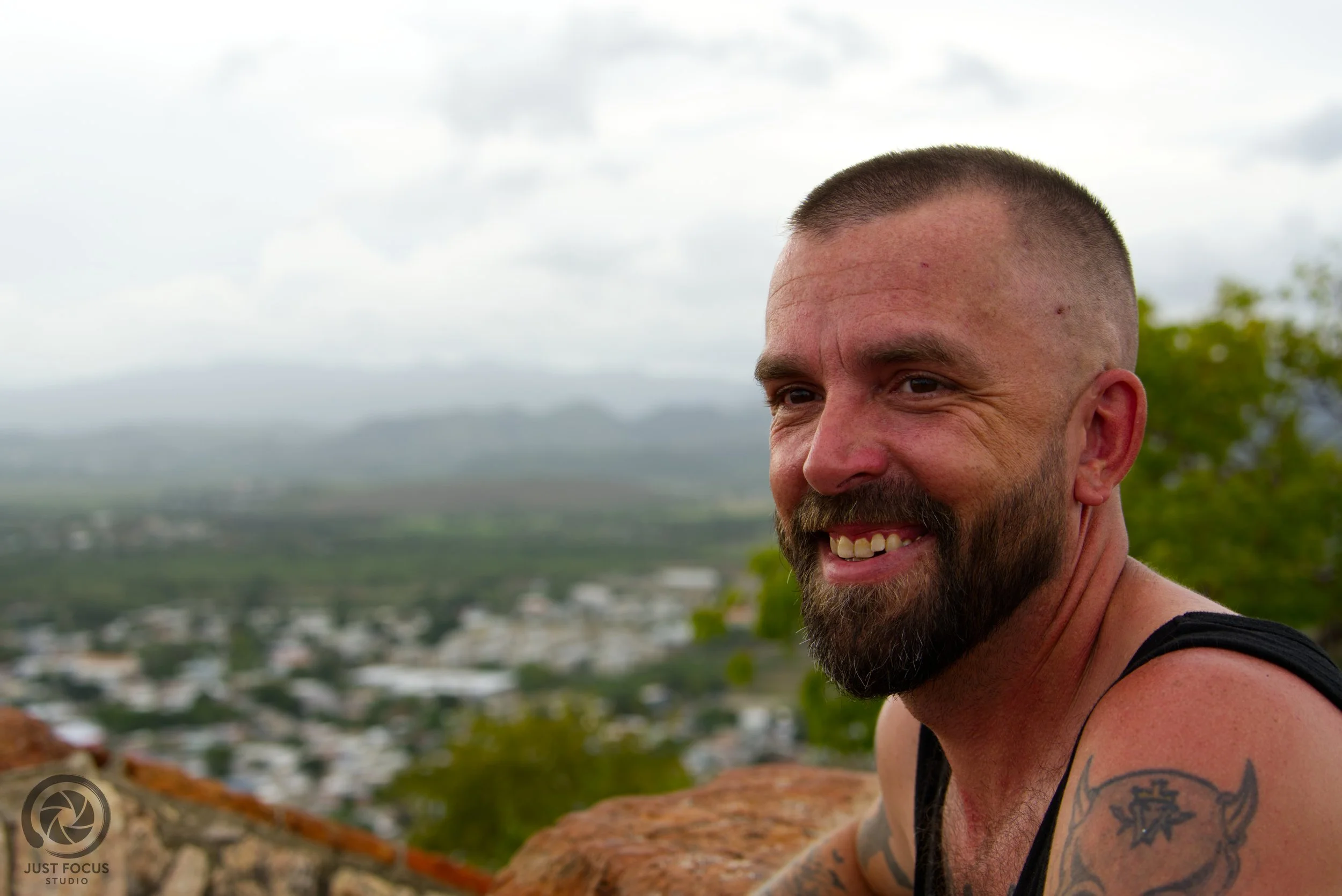 Close-up of a smiling man with a beard and tattooed arm, outdoors with a blurred landscape of a city and mountains in the background.
