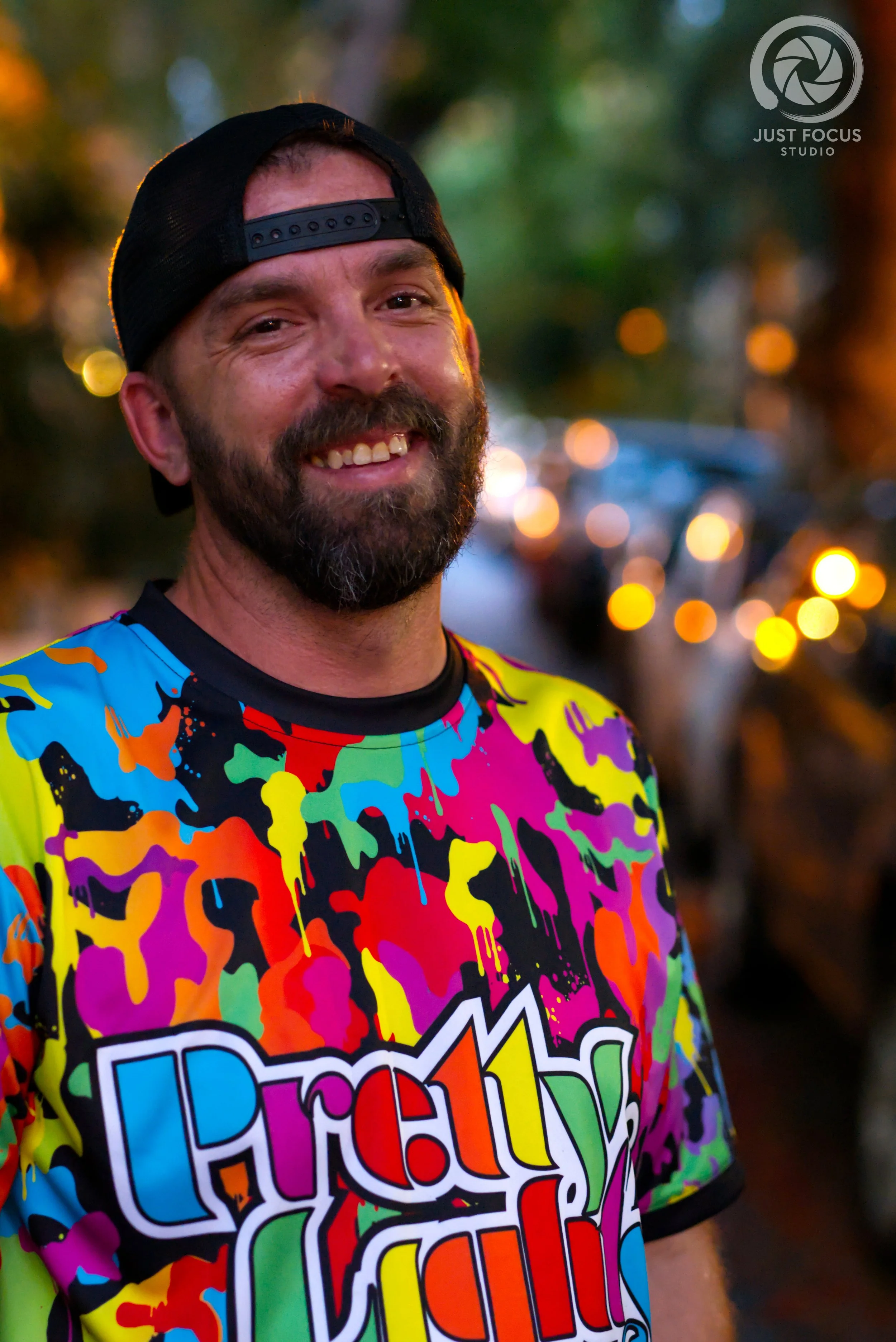A smiling man with a beard, wearing a colorful, splatter-patterned t-shirt and a black backwards cap, standing outdoors during dusk with blurred lights in the background.
