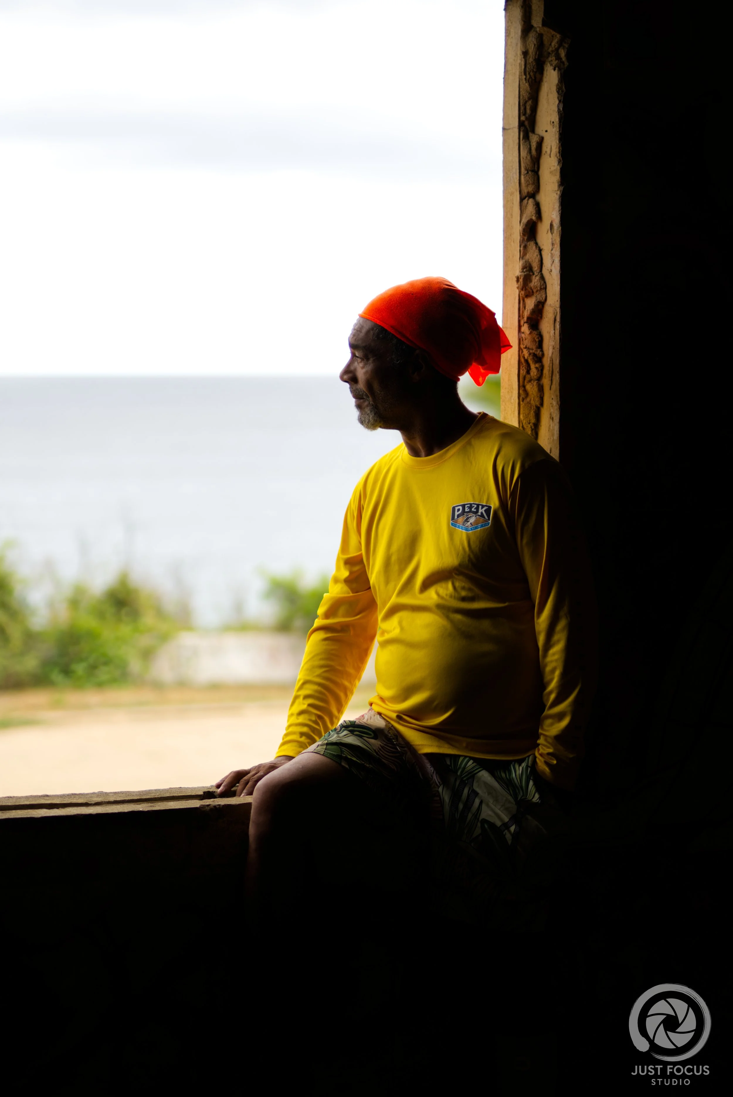 Silhouette of an older man sitting by a window looking outside, wearing a red headscarf and yellow shirt, with a blurred outdoor background.