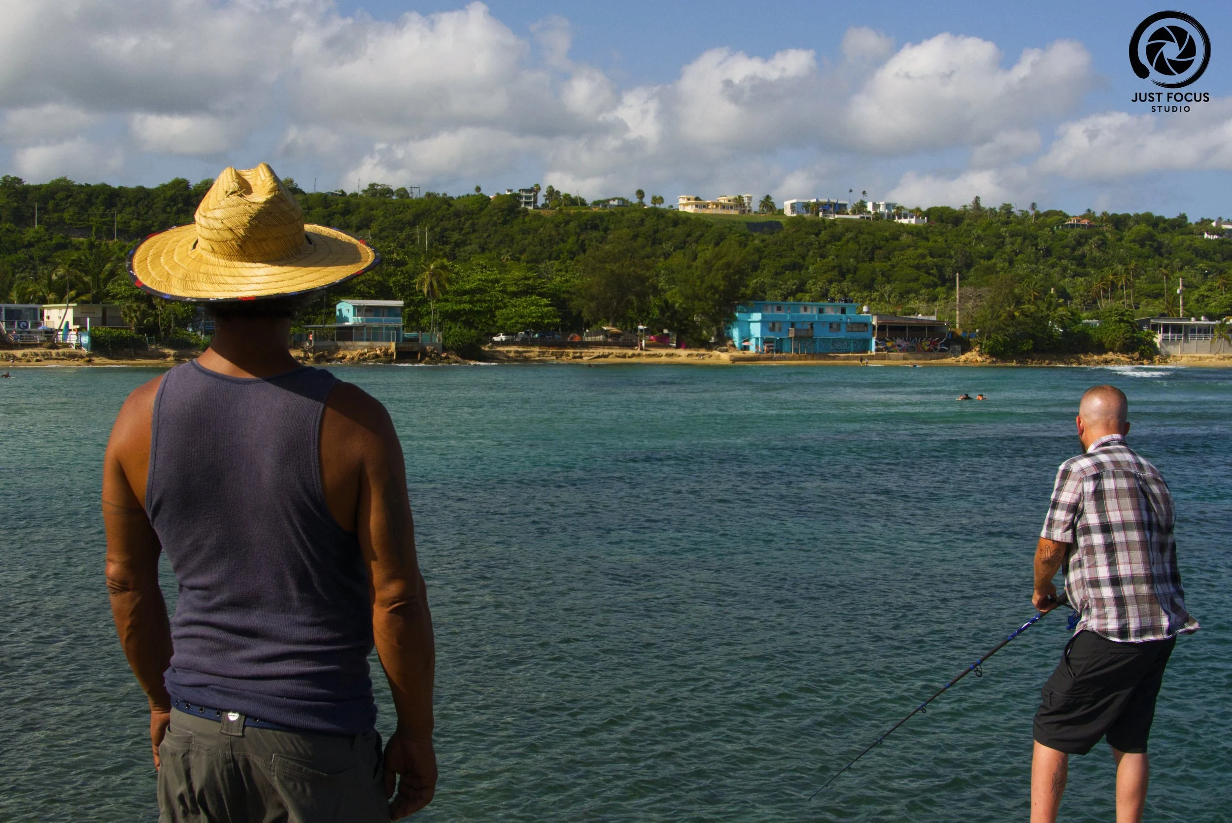 Two men standing by the water side; one wearing a wide-brimmed straw hat and a sleeveless shirt, the other wearing a short-sleeved plaid shirt and shorts, fishing in a coastal area with houses and greenery in the background under a partly cloudy sky.