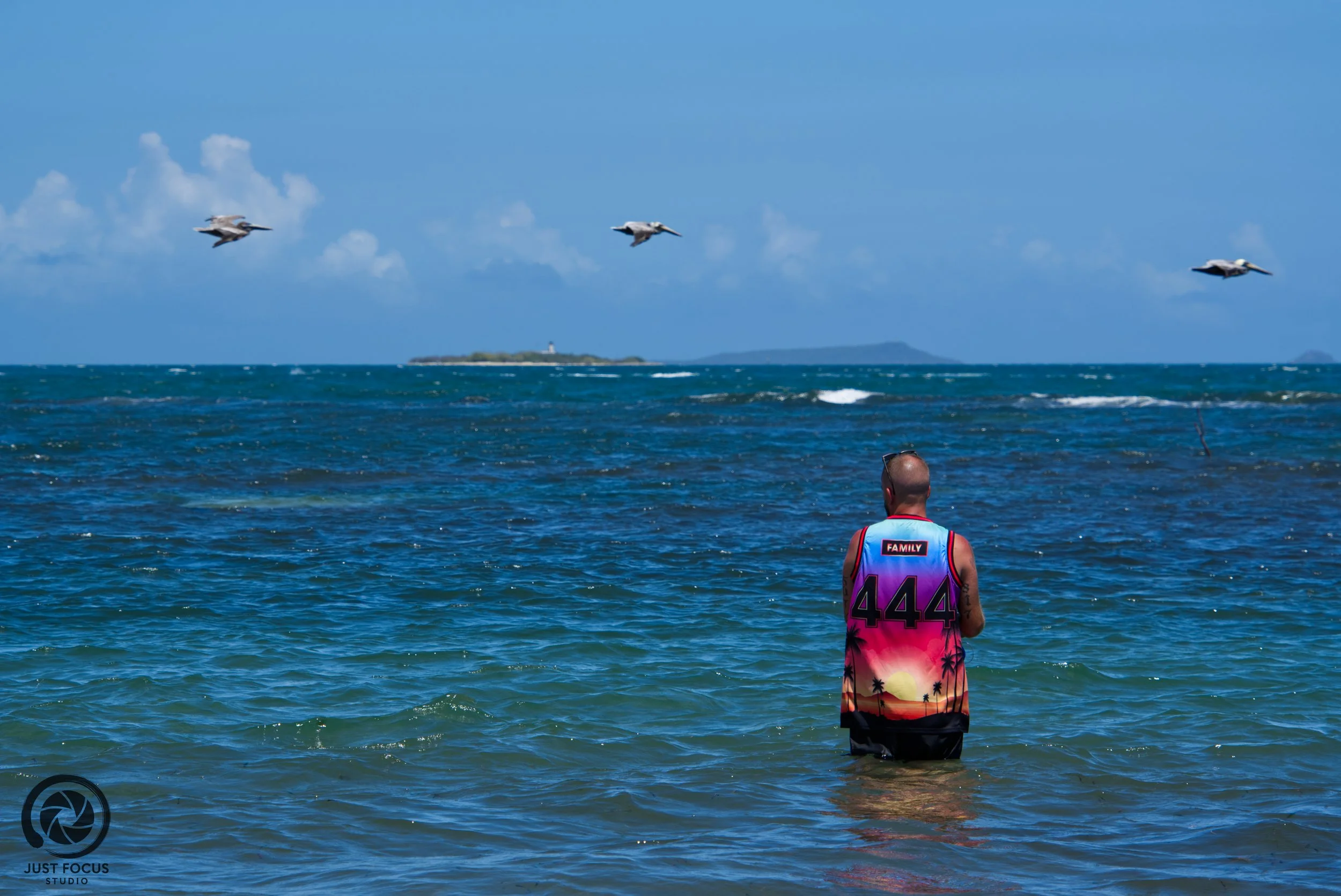 A person standing in the ocean, facing away, wearing a colorful sleeveless shirt with the number 444 and a sunset scene, with three planes flying overhead and a distant landmass on the horizon.