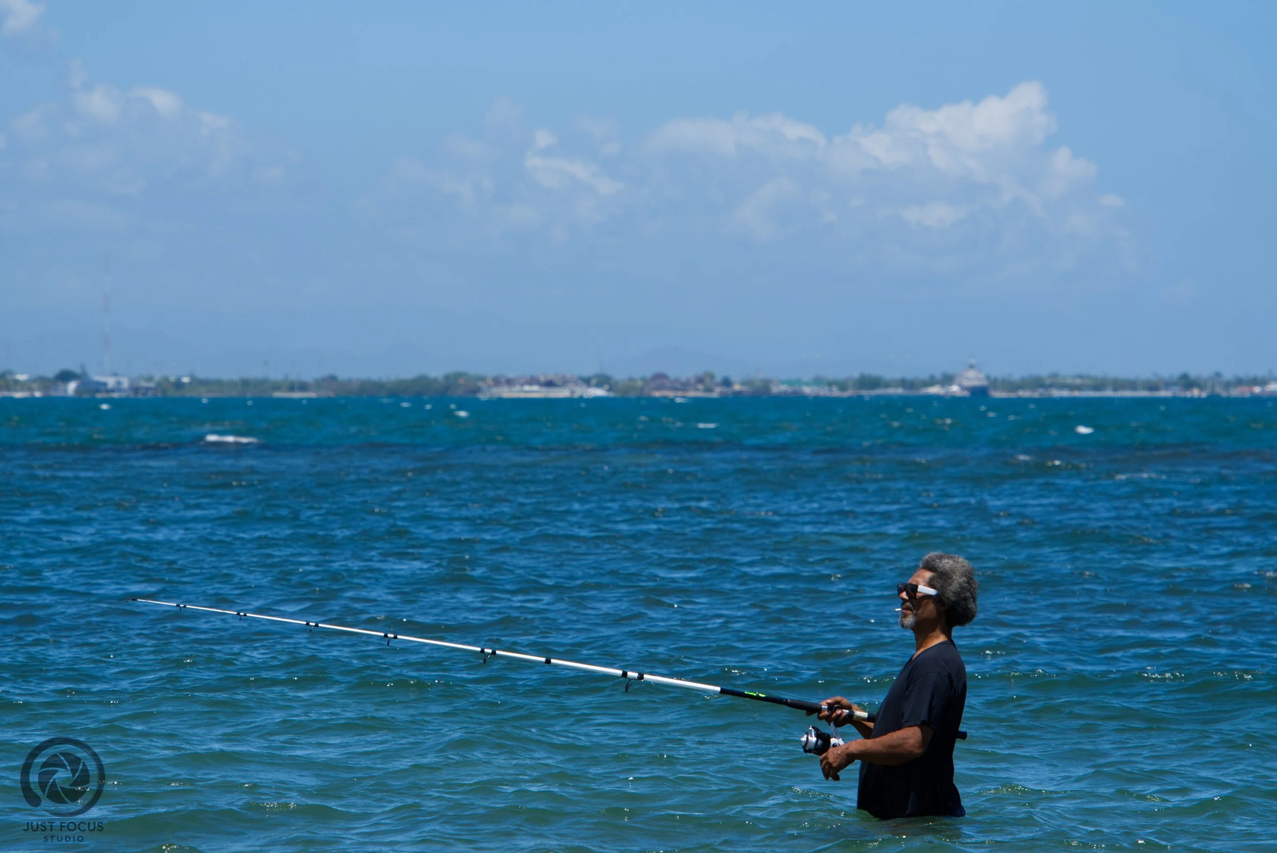 A man standing in the ocean holding a fishing rod, wearing sunglasses and a black shirt, with a city skyline and blue sky with clouds in the background.
