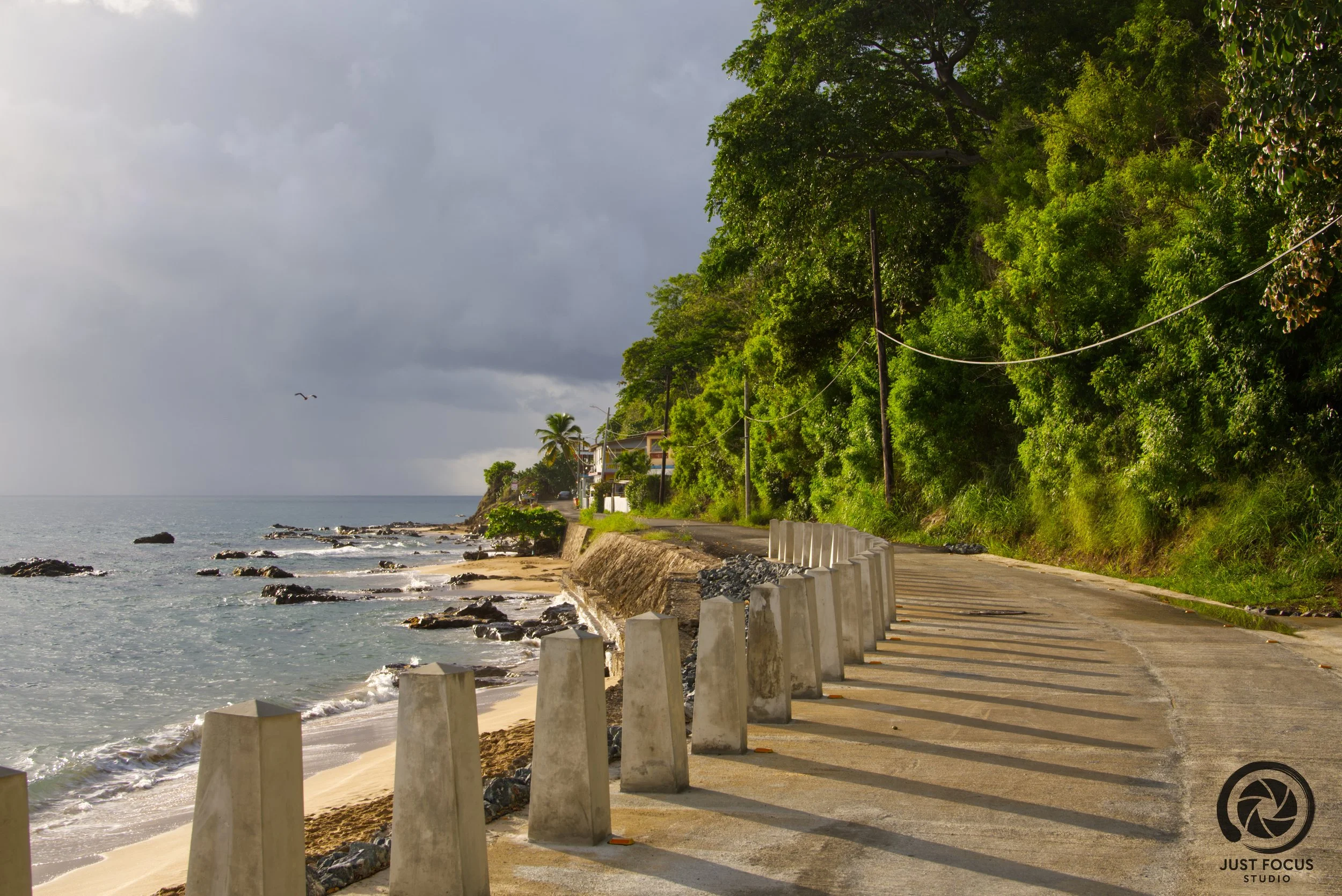 A coastal road with concrete barriers on the beach side and lush green trees on the other side. The ocean is visible to the left with some rocks and a cloudy sky overhead.