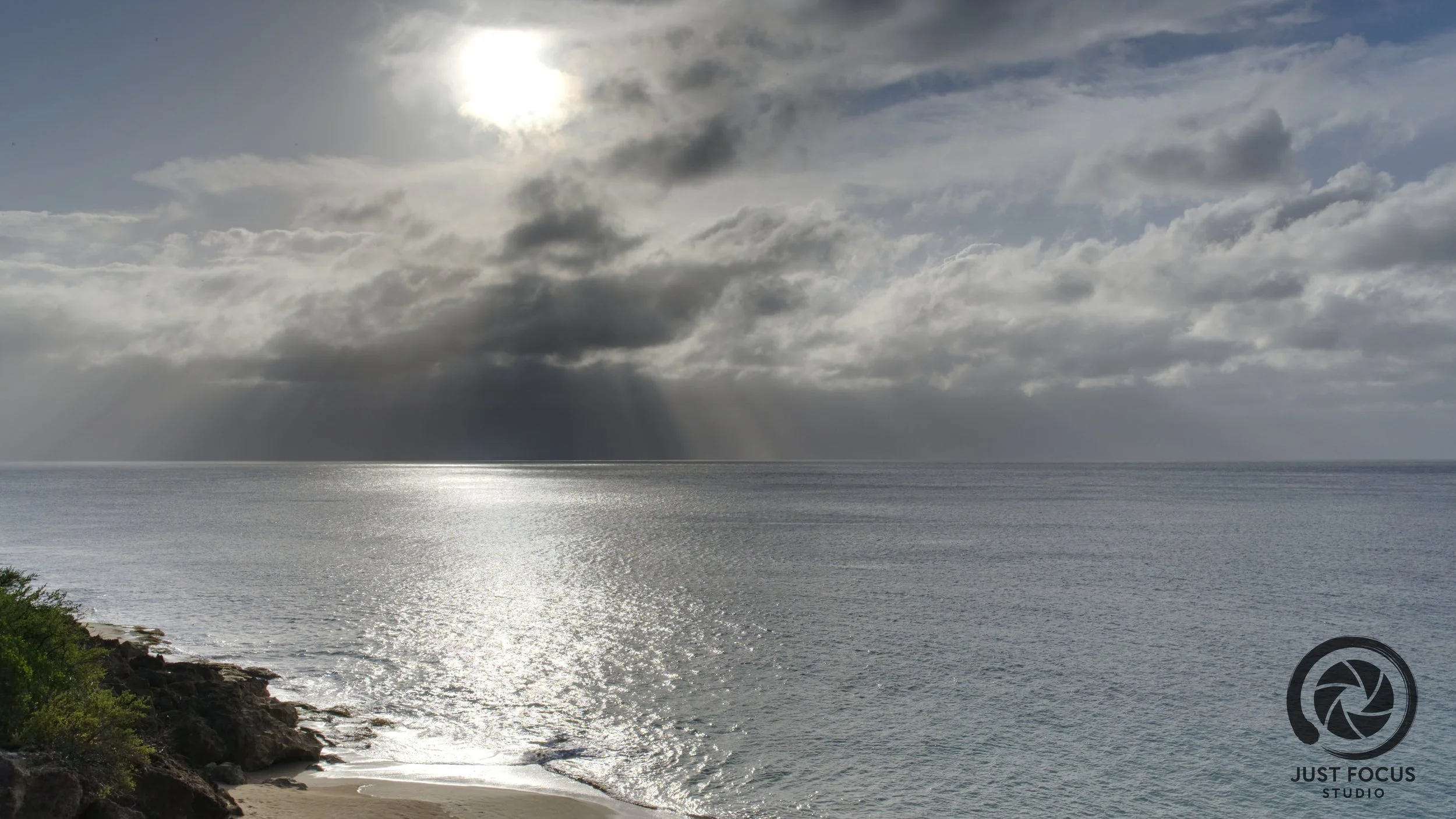 Overcast sky over a calm ocean with sunlight shining through clouds, rocky shoreline in the foreground, and a logo for Just Focus Studio in the lower right corner.