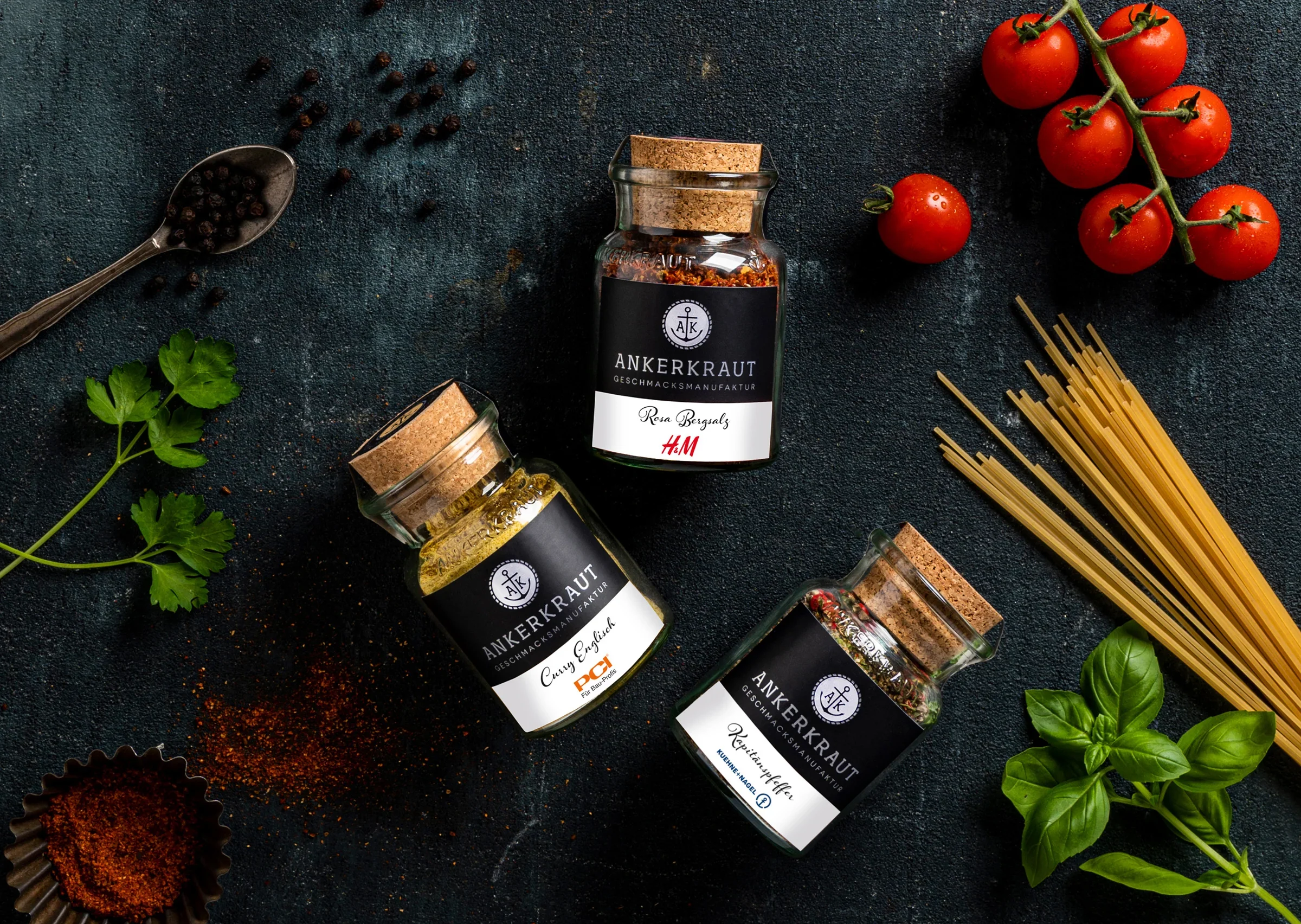 Drei Gläser mit Gewürzen, darunter Ankerkraut-Sorten, befinden sich auf einem schwarzen Oberflächenhintergrund. Es gibt auch frische Tomaten, ein Bund Basilikum, Pasta und Gewürzpaprika in einem kleinen Förmchen.