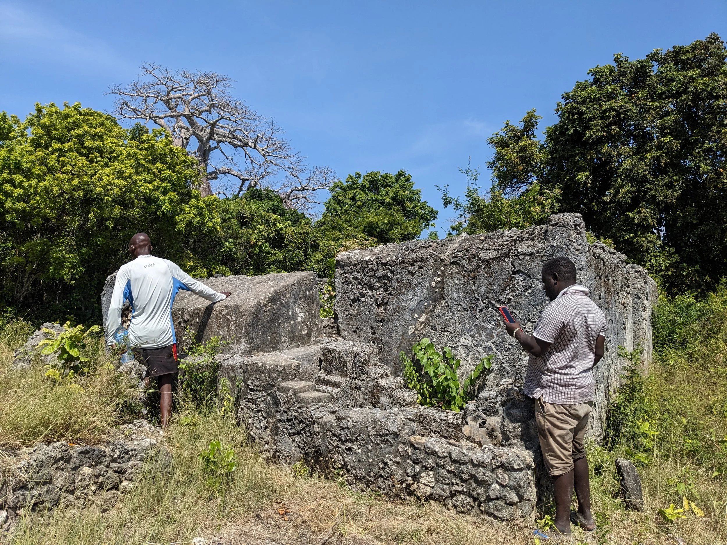 Two men stand near ancient stone ruins surrounded by greenery, with a clear blue sky overhead.