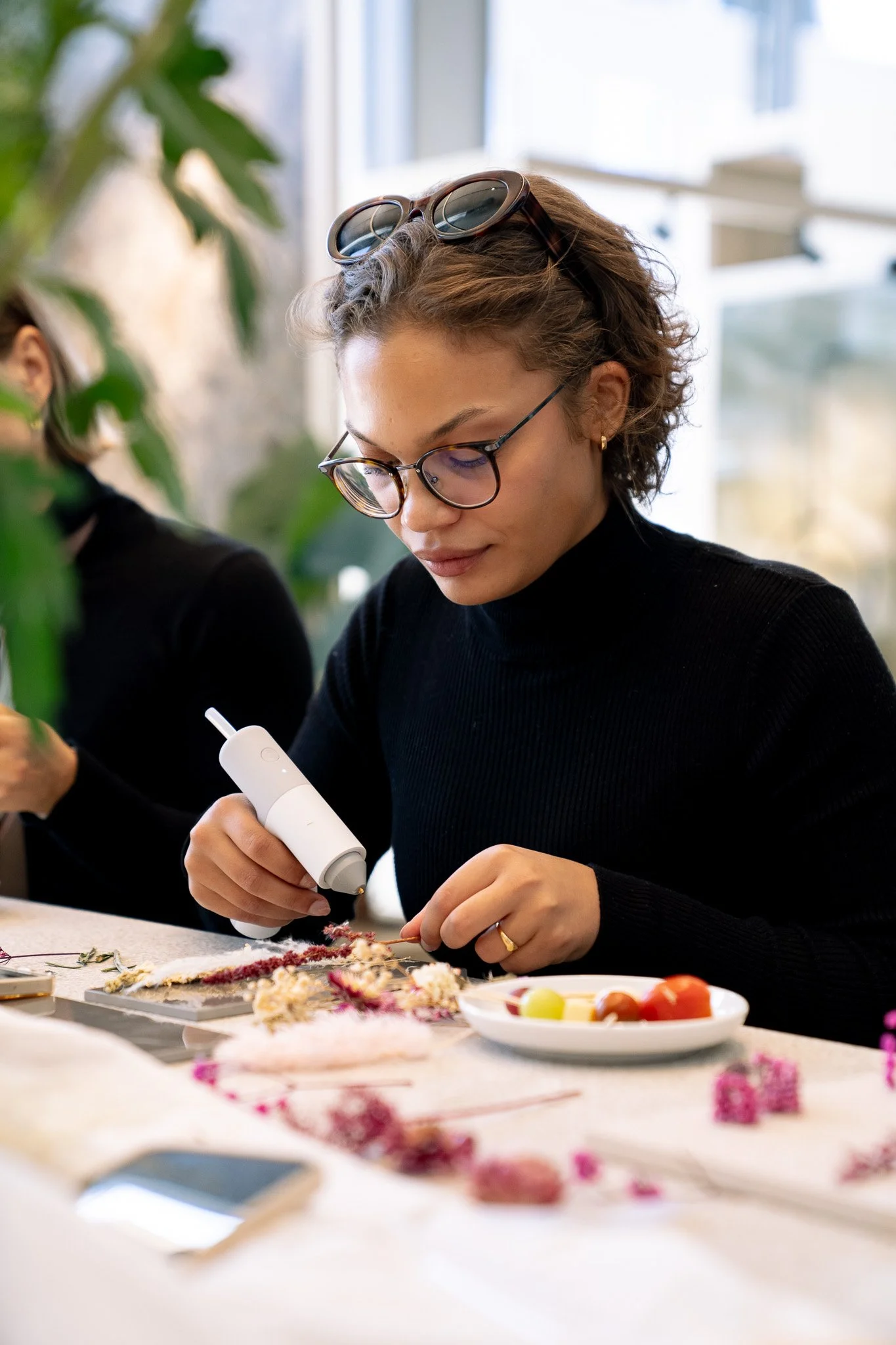 Frau beim Basteln von Blumen mit Heißklebepistole, auf dem Tisch sind bunte Blumen, Steine und Obst.