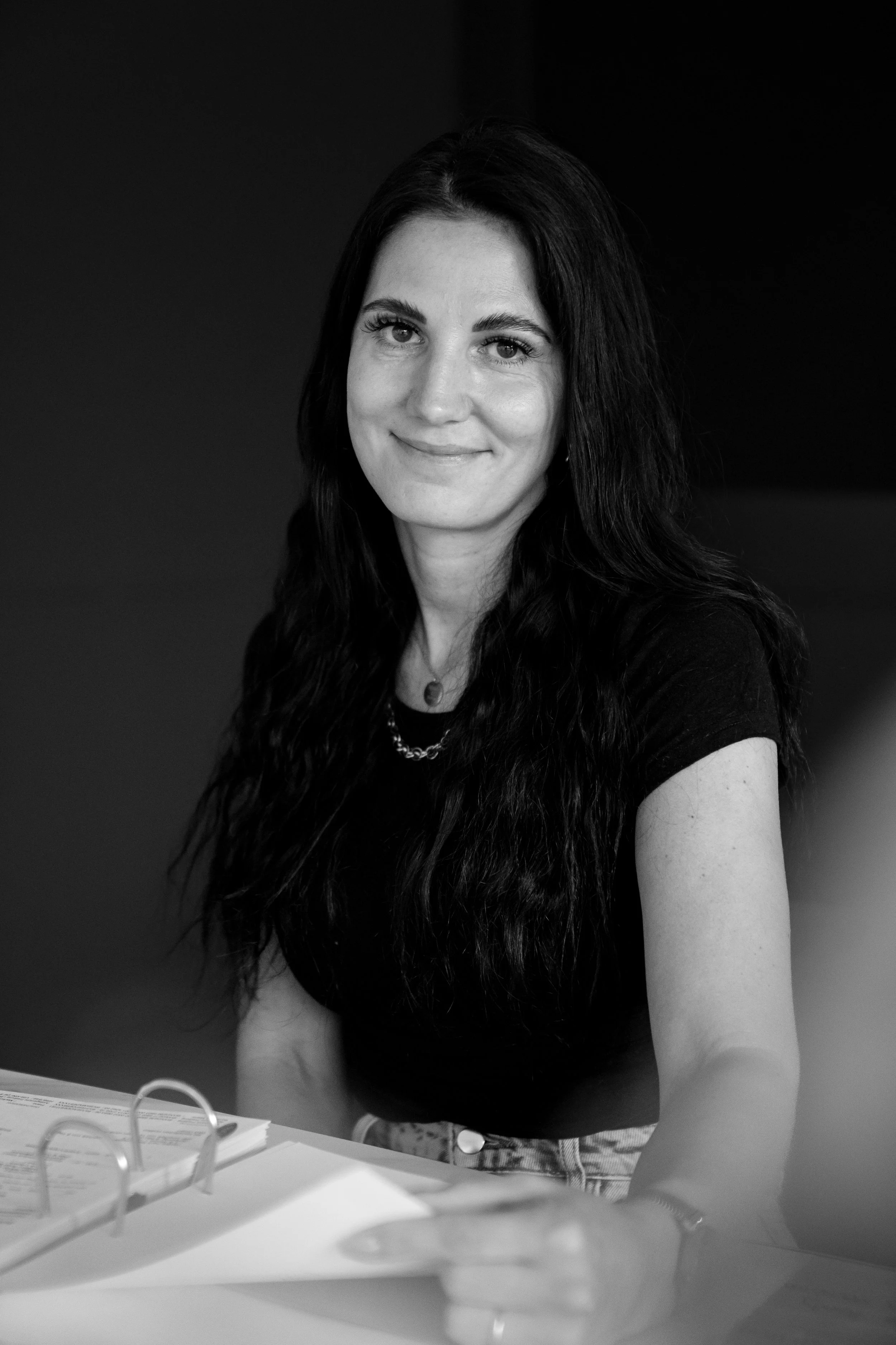 Schwarz-weiß Porträt einer jungen Frau mit langen dunklen Haaren, lächelnd, sitzend an einem Tisch, schaut in die Kamera.
