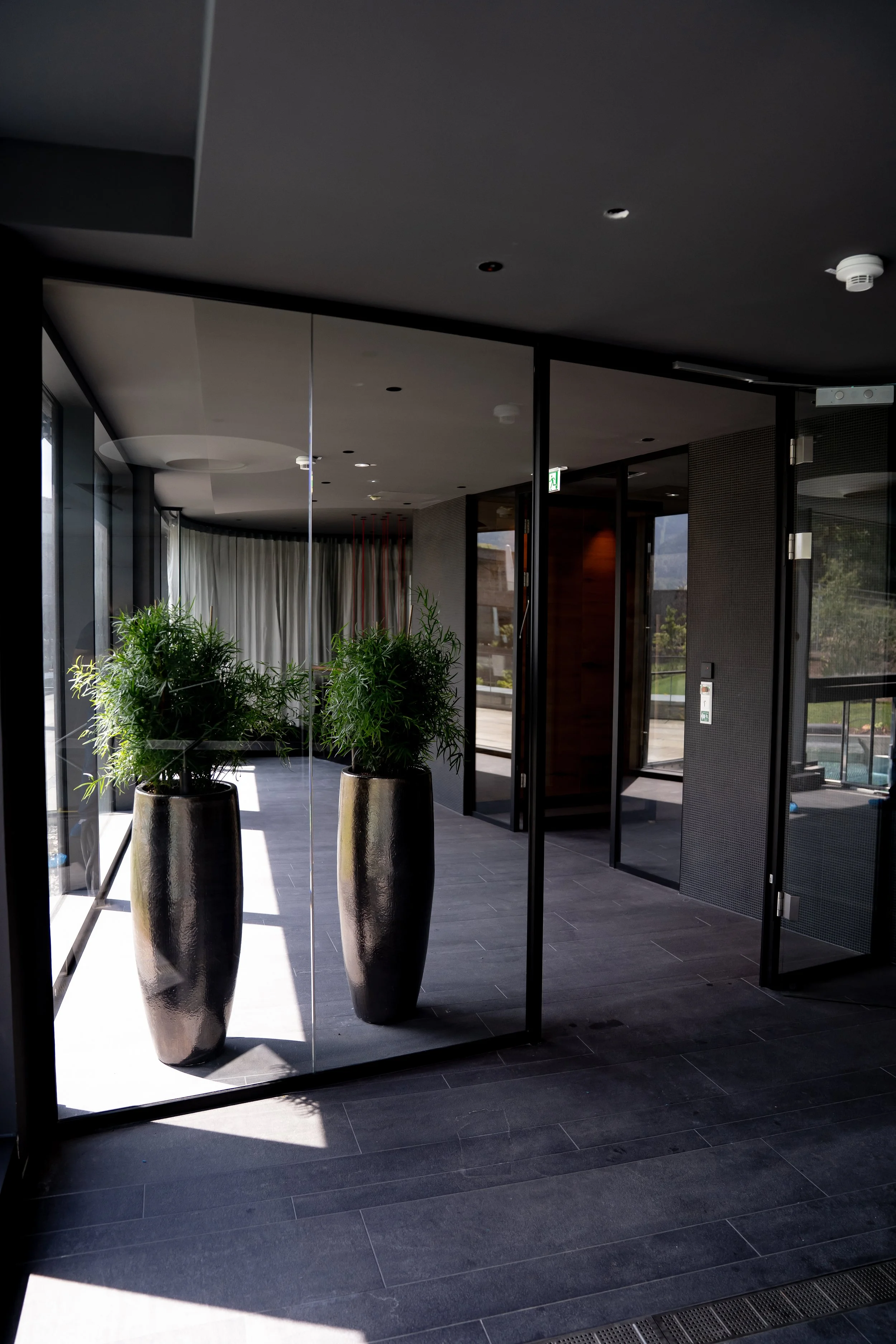 Innenaufnahme eines modernen Gebäudes mit zwei großen schwarzen Vasen und grünen Pflanzen, glasartige Türen und Fenster, dunkle Fliesenboden, natürlicher Lichtschein.