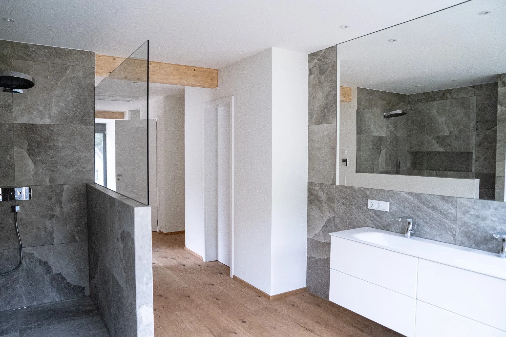 Moderner Badezimmerraum mit grauen Fliesen, weißem Waschtischunterschrank, großen Spiegel und Holzfußboden.