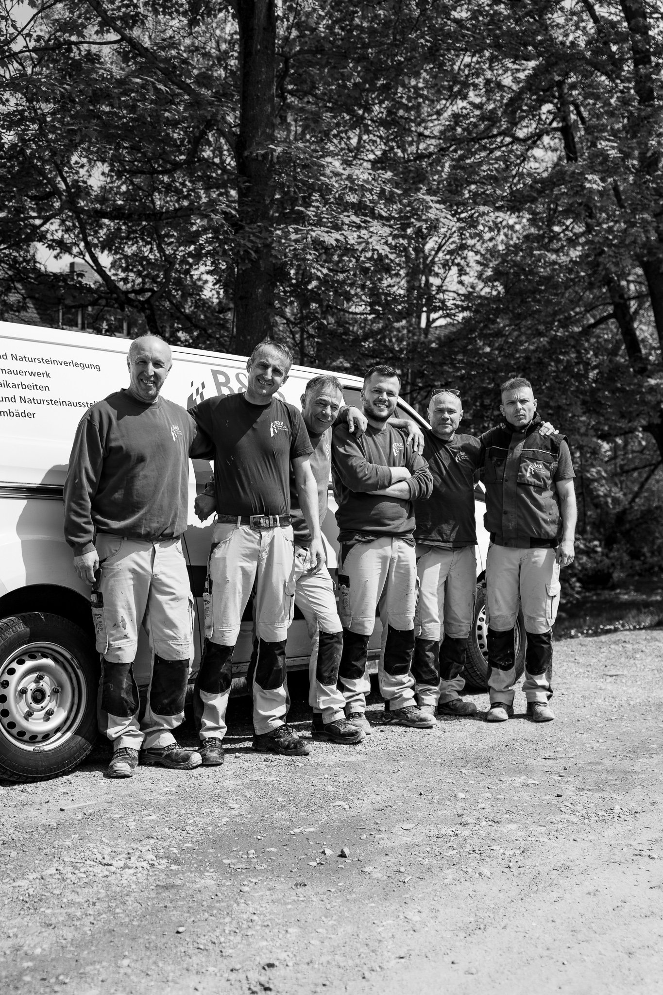 Fünf Männer in Arbeitshosen und Oberteilen stehen nebeneinander vor einem weißen Dienstwagen im Freien, umgeben von Bäumen, lächelnd in die Kamera blickend.